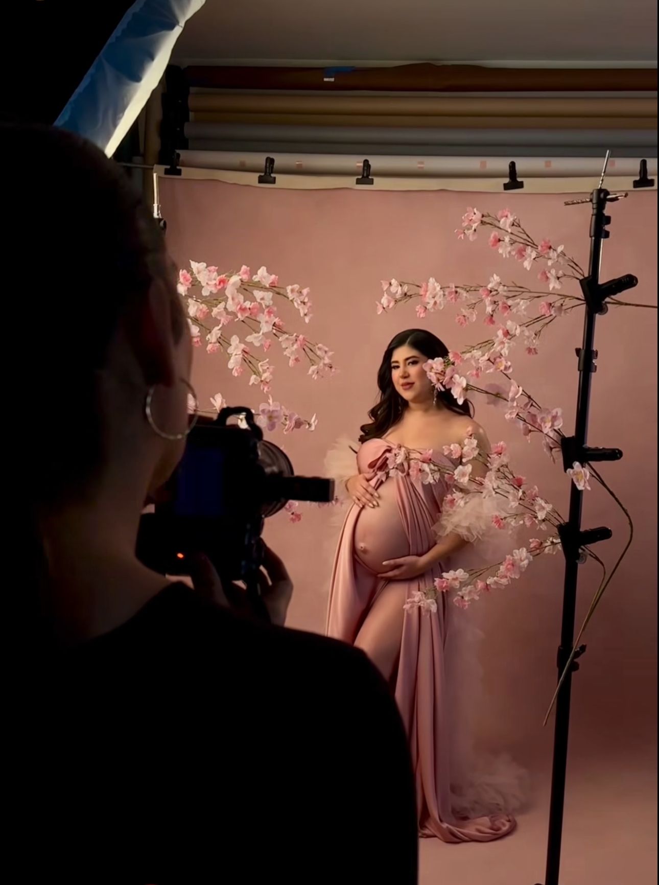 Photographer captures pregnant woman in pink gown posing before cherry blossom arch in professional studio setup.