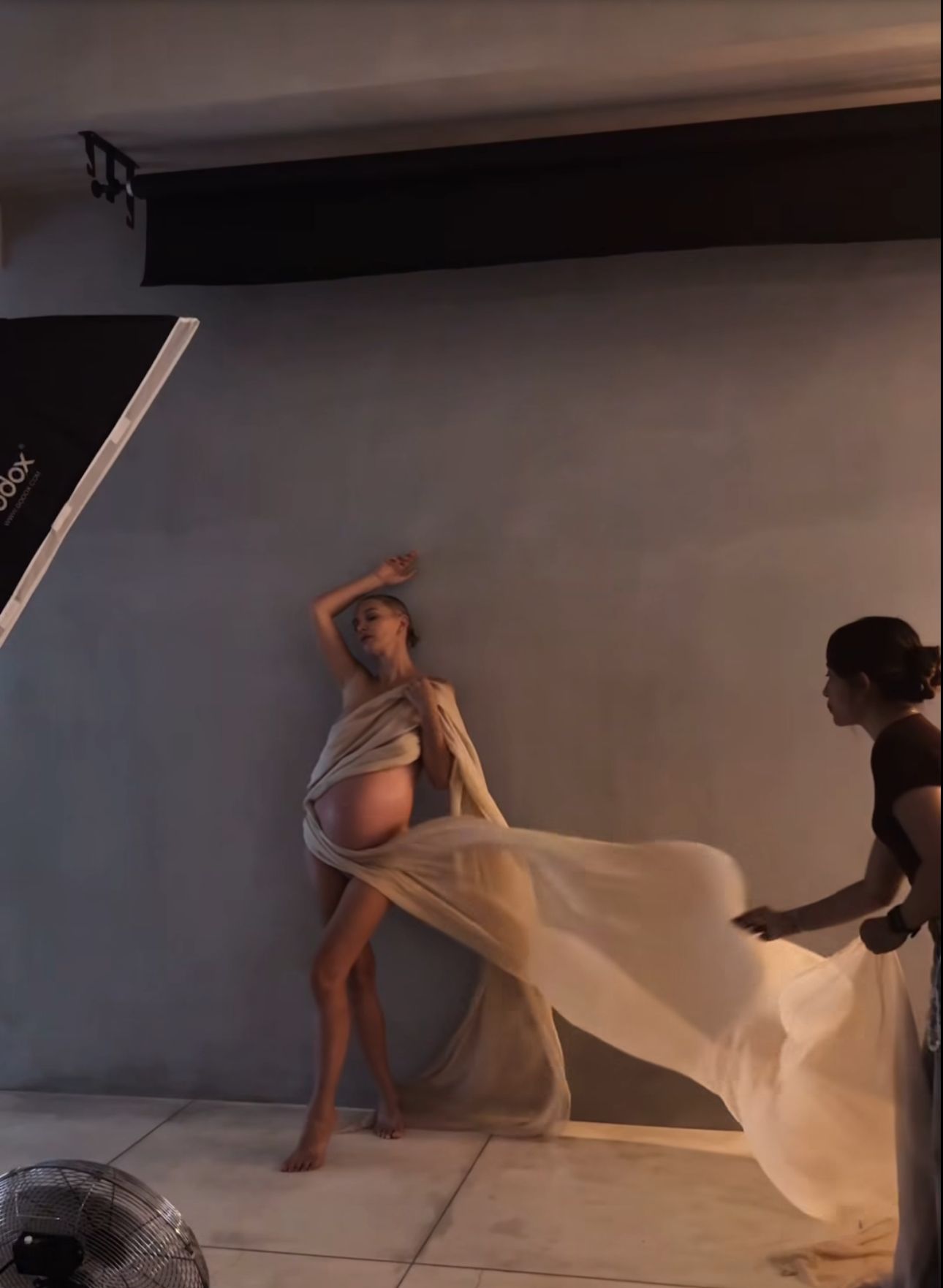 Pregnant woman in flowing sheer fabric poses during a maternity photoshoot in a studio setting.