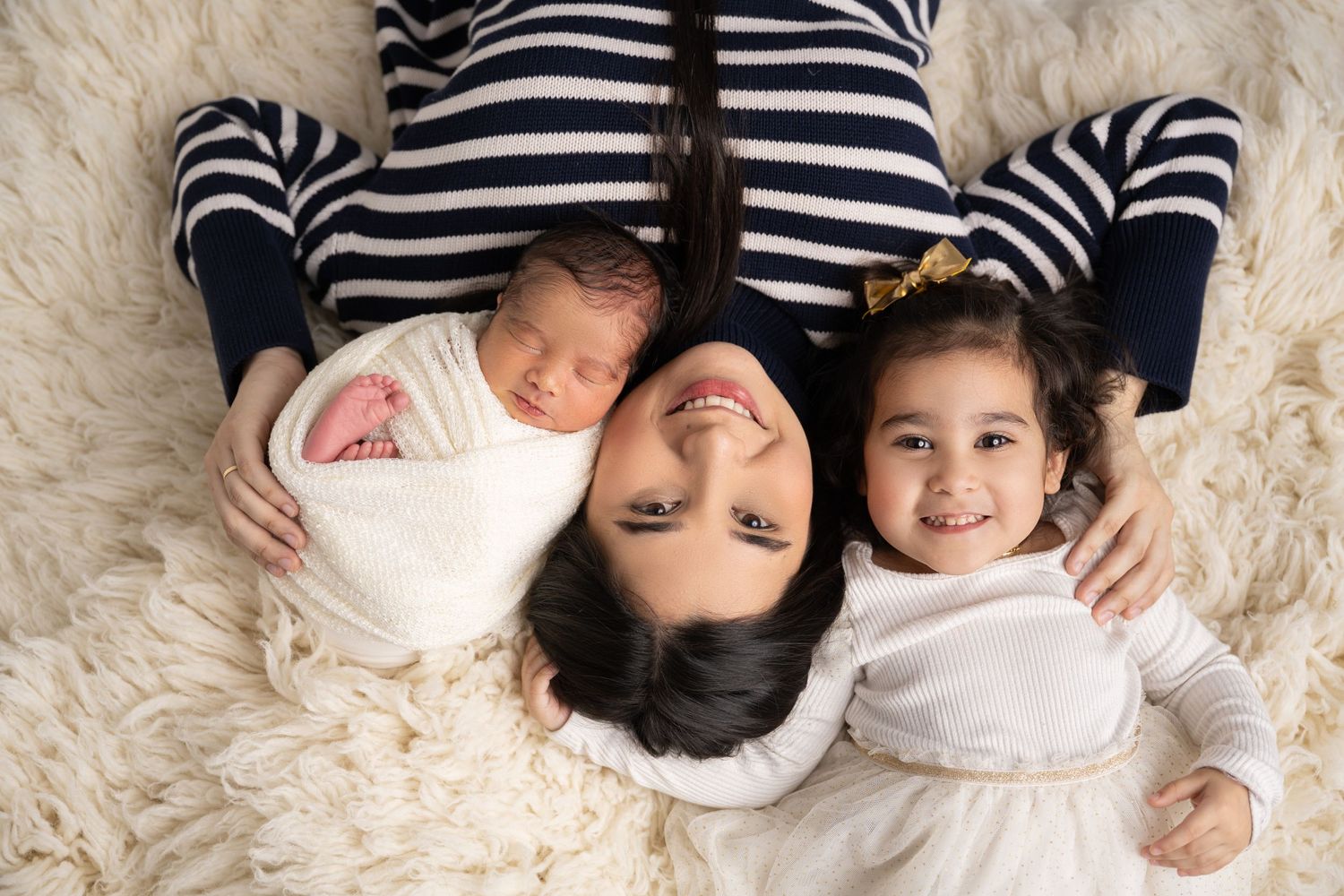 Family portrait on cream fur rug with newborn and siblings in coordinated white and navy outfits.