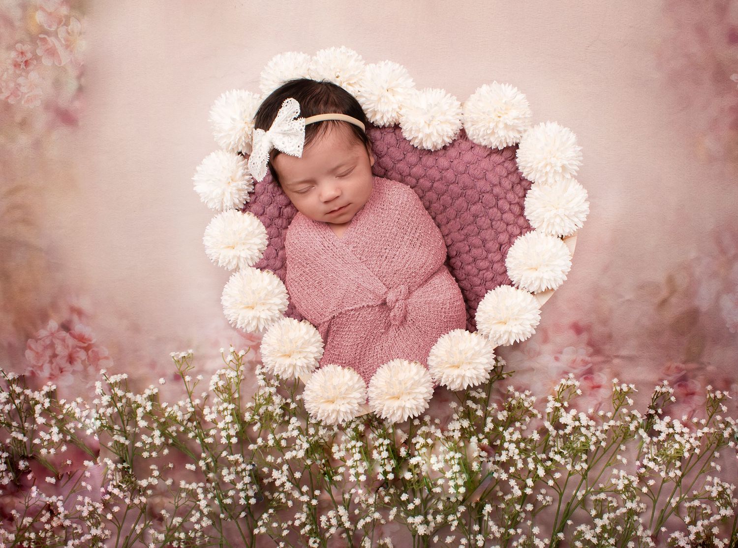 Sleeping newborn in pink outfit surrounded by white floral heart shape and baby's breath flowers.