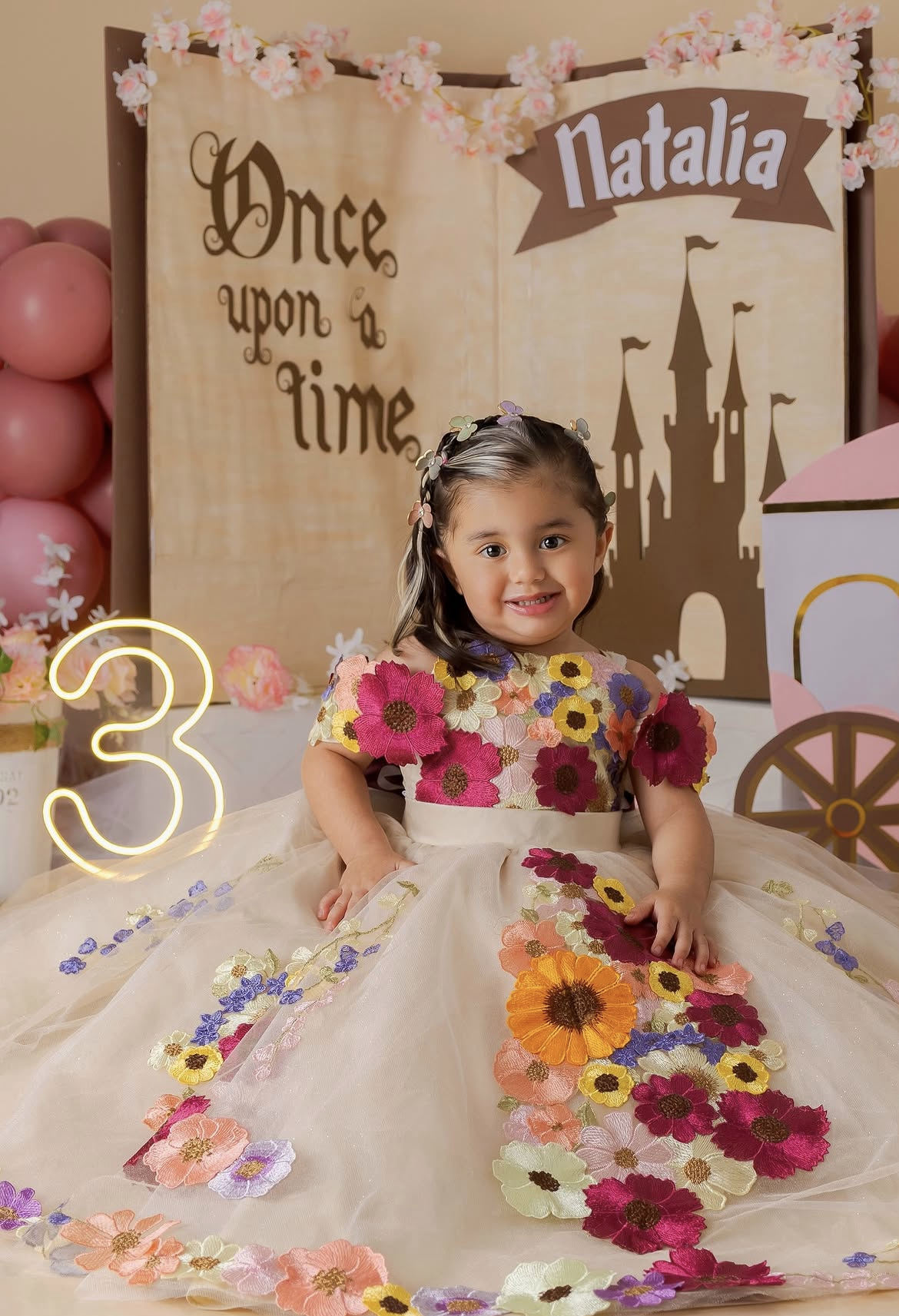 Girl in floral dress celebrates 3rd birthday with Once Upon a Time princess theme backdrop and name sign Natalia.