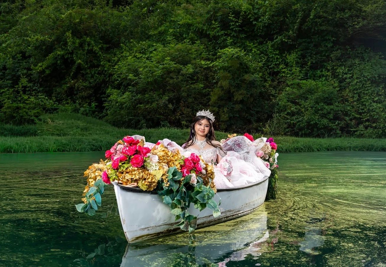 A woman in a floral crown sits in a flower-filled white boat on a serene green lake surrounded by lush trees.