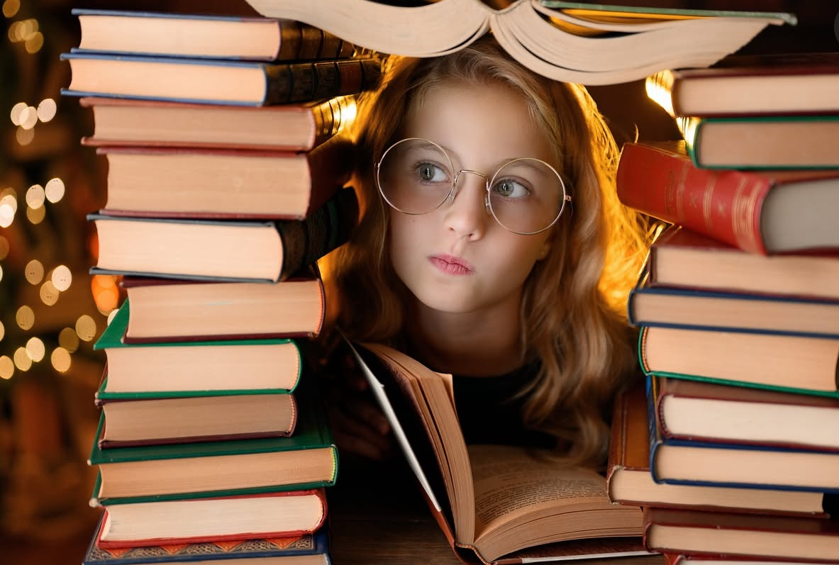 A young girl with glasses reads a book surrounded by tall stacks of colorful books in a warm, cozy setting.