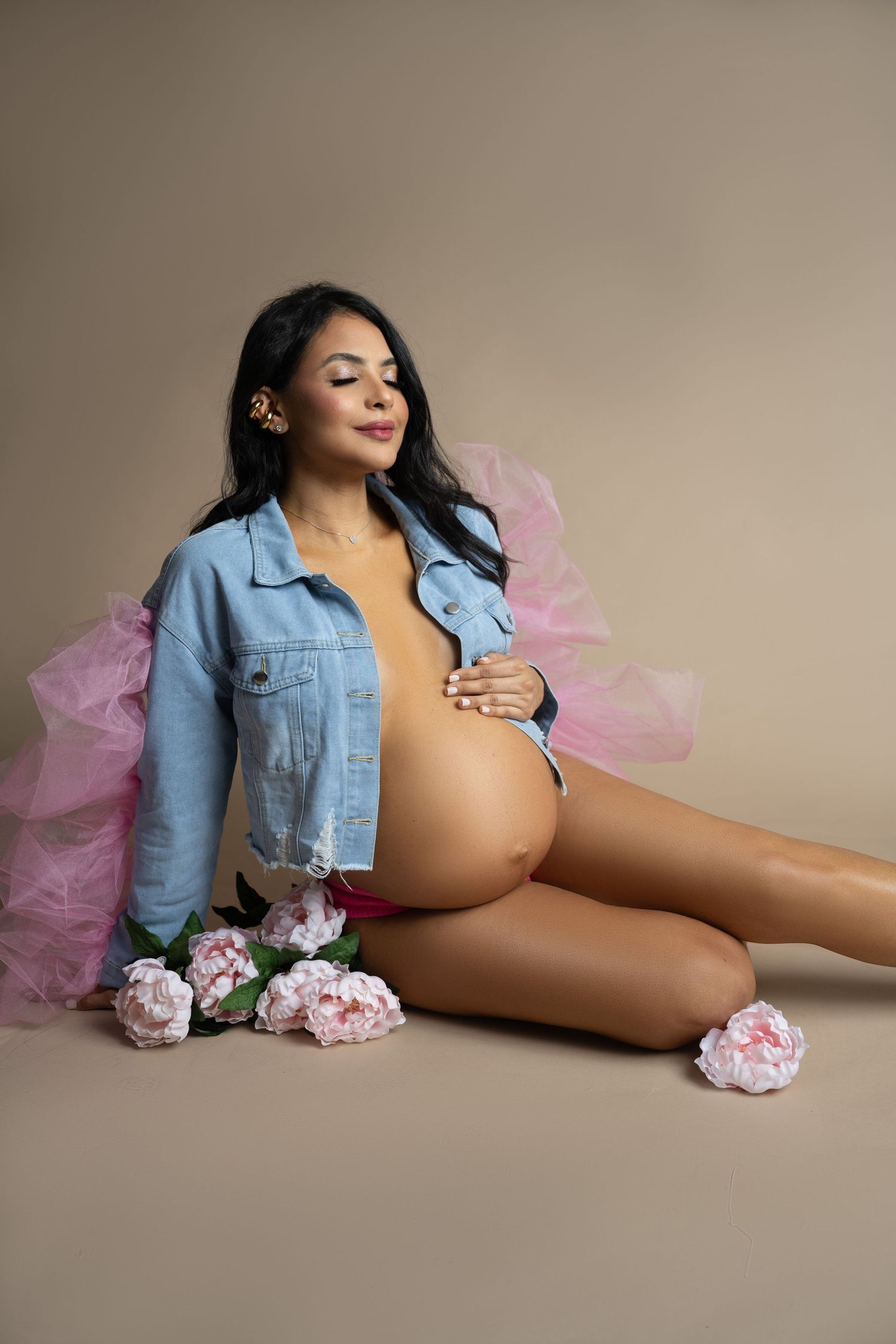 Maternity portrait with denim jacket and pink flowers against neutral background.