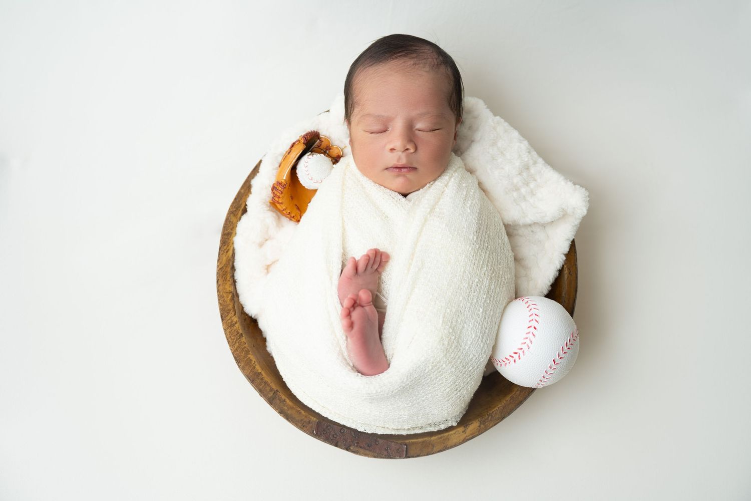 Newborn wrapped in white blanket sleeping peacefully in round wooden bowl.
