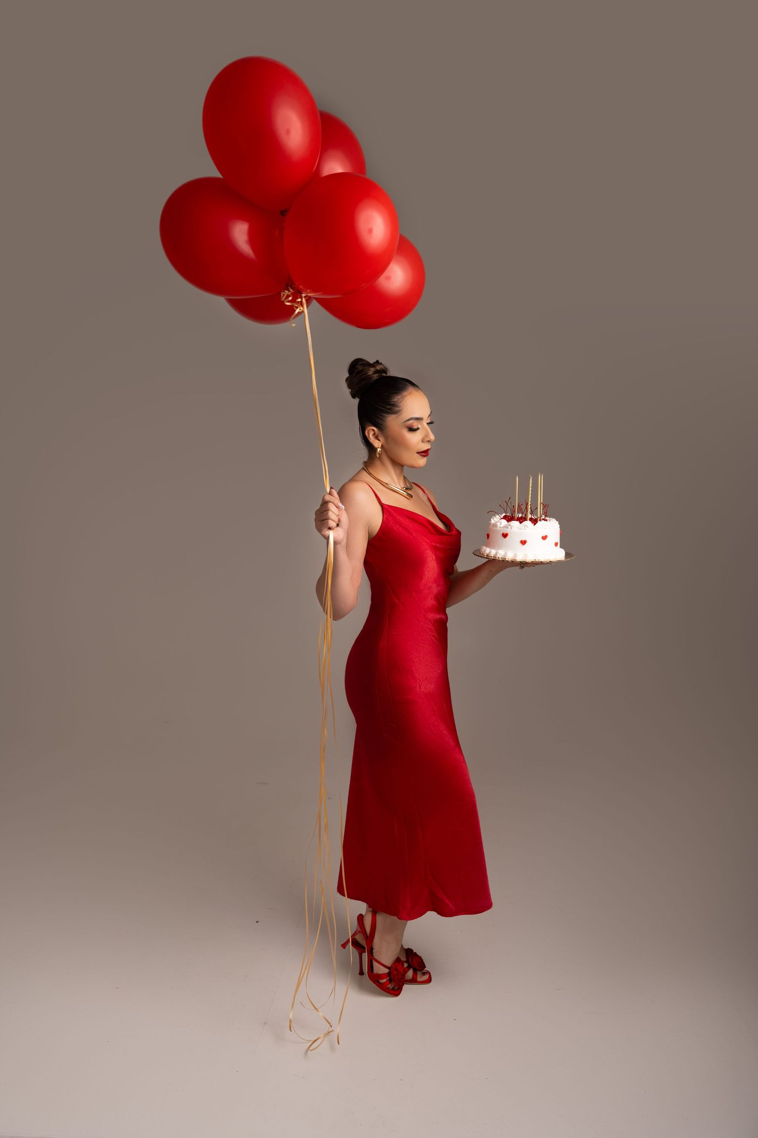 Elegant figure in red dress holds red balloons and birthday cake while posing against neutral background.