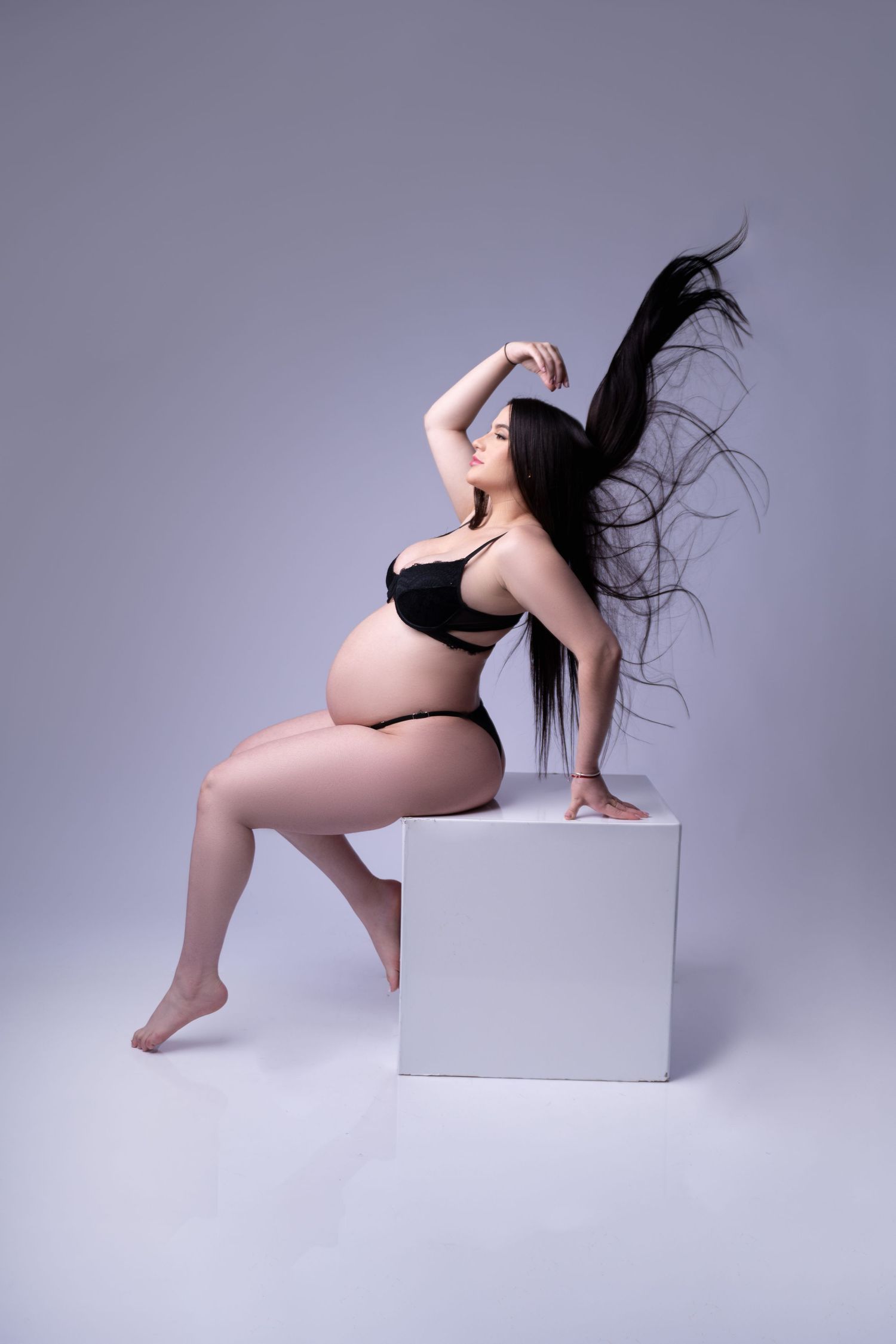 Dynamic maternity pose with flowing hair on white cube against grey background.