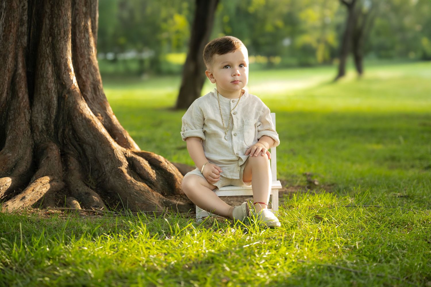 A child sits in white clothing beneath a large tree trunk on grass during a sunny day.