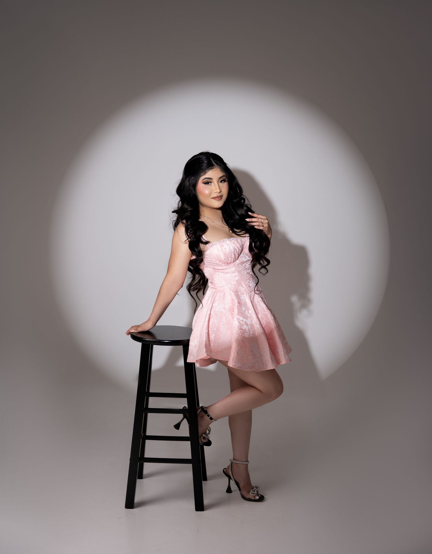Young woman in pink mini dress posing beside black stool against white spotlight backdrop in studio.