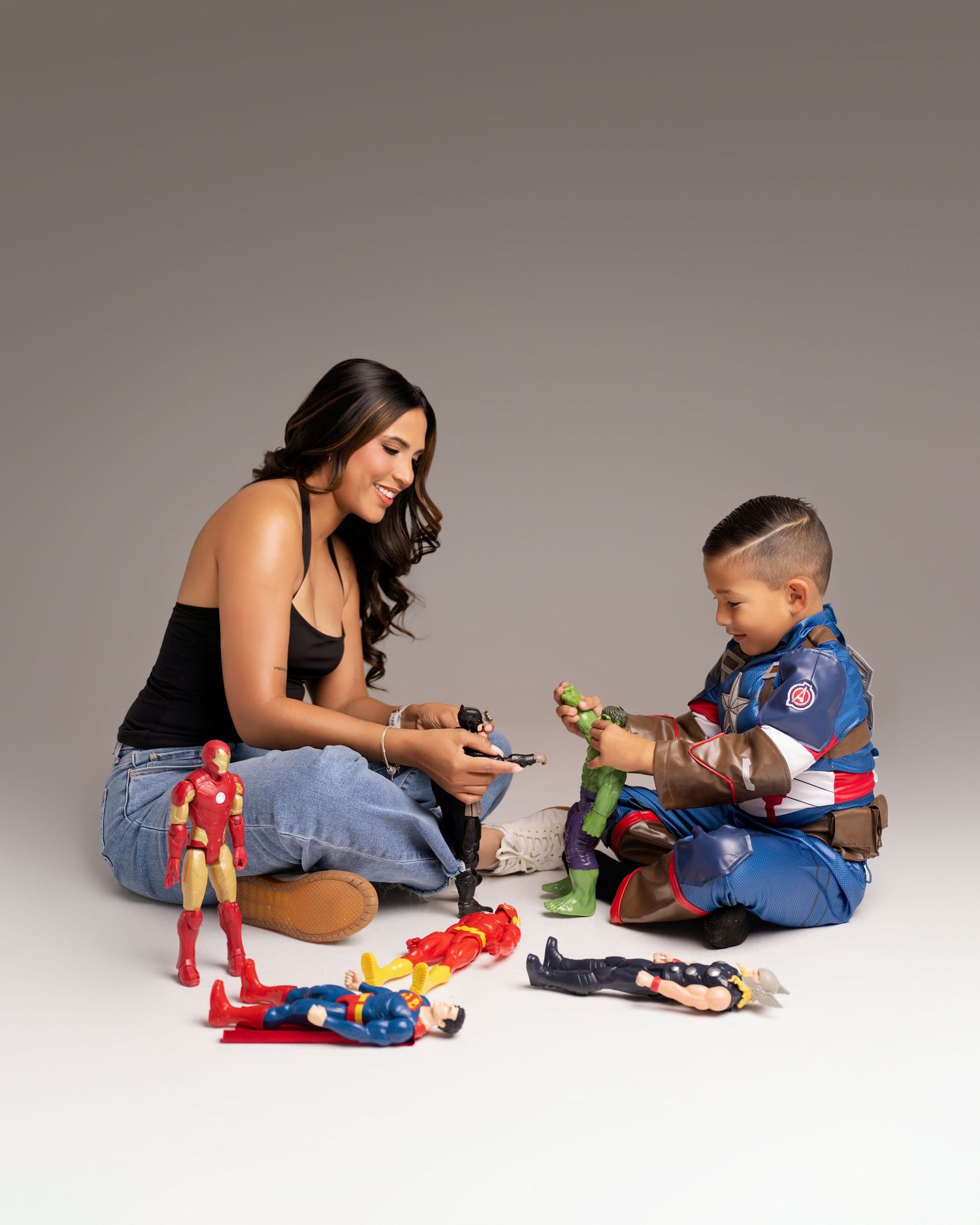 A woman and young boy in a Captain America costume play with Marvel superhero action figures on a studio floor.