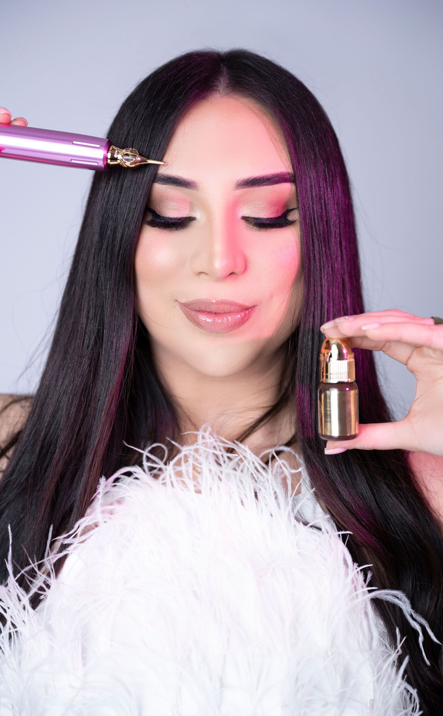 Woman with long dark hair and pink highlights applies gold serum with pink applicator wand, wearing white feather top.