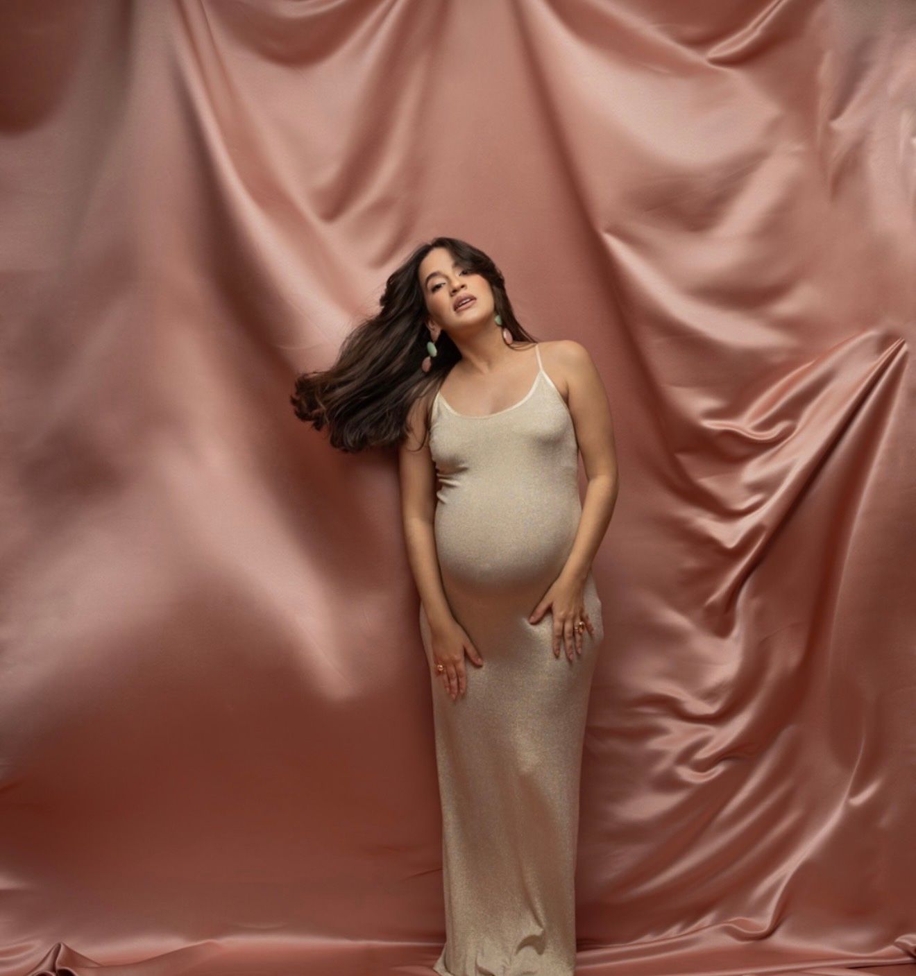 Pregnant woman in elegant cream slip dress posing gracefully against pink satin backdrop.
