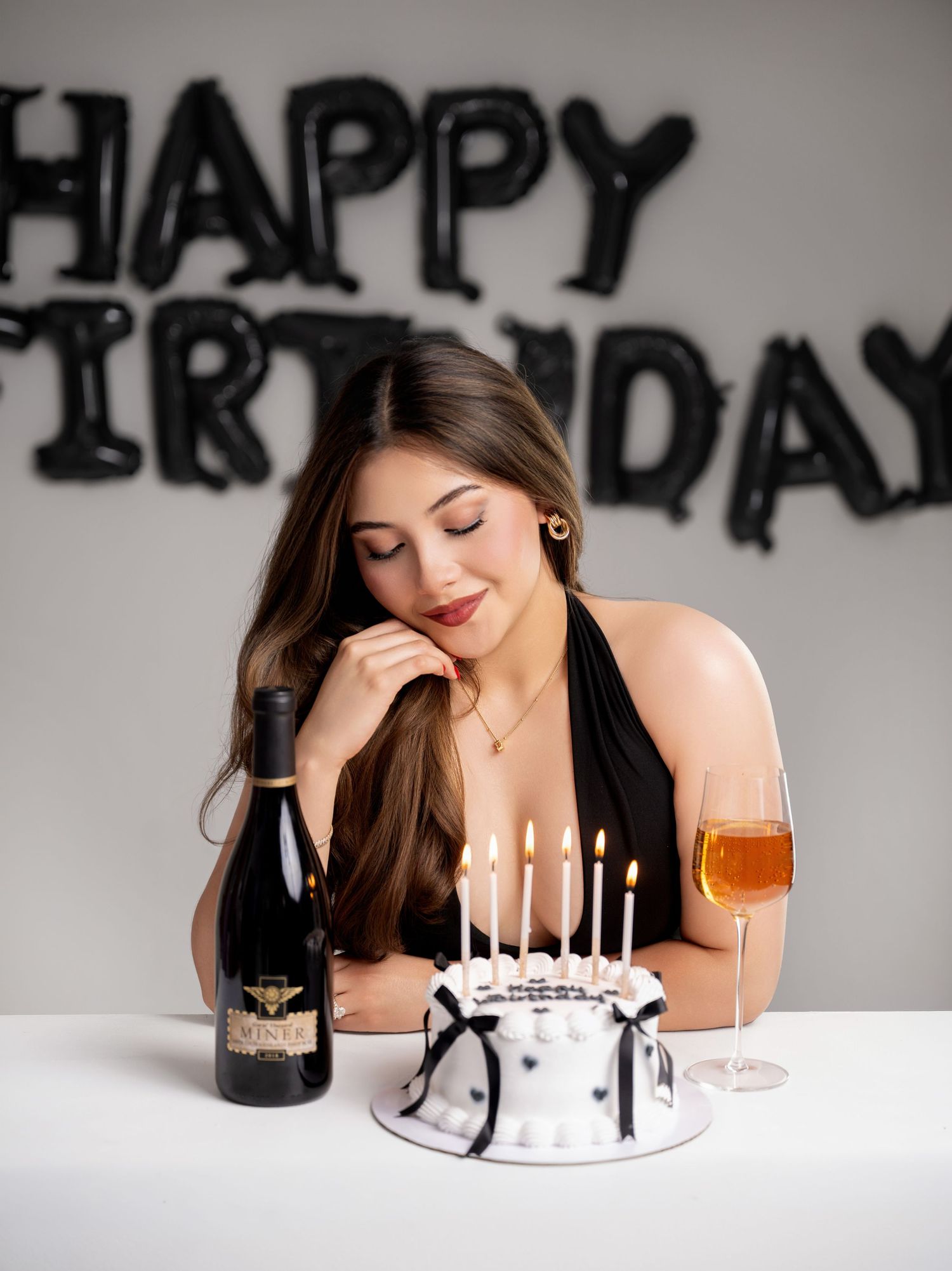 Woman with long brown hair makes a wish over a lit white birthday cake beside champagne and a bottle, with Happy Birthday balloons behind.