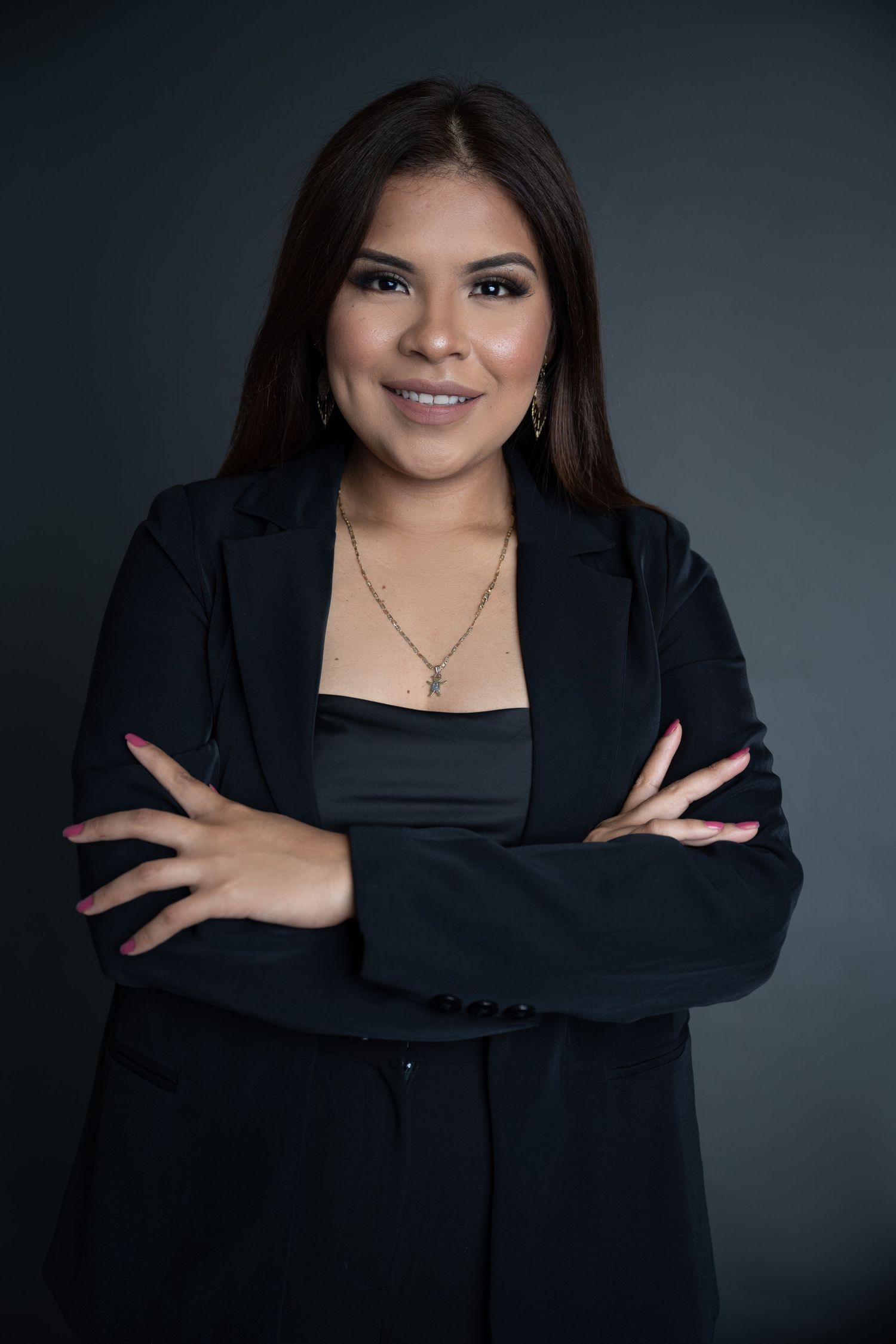 Smiling woman with long dark hair, wearing a black blazer and pendant necklace, poses with arms crossed against a dark studio backdrop.