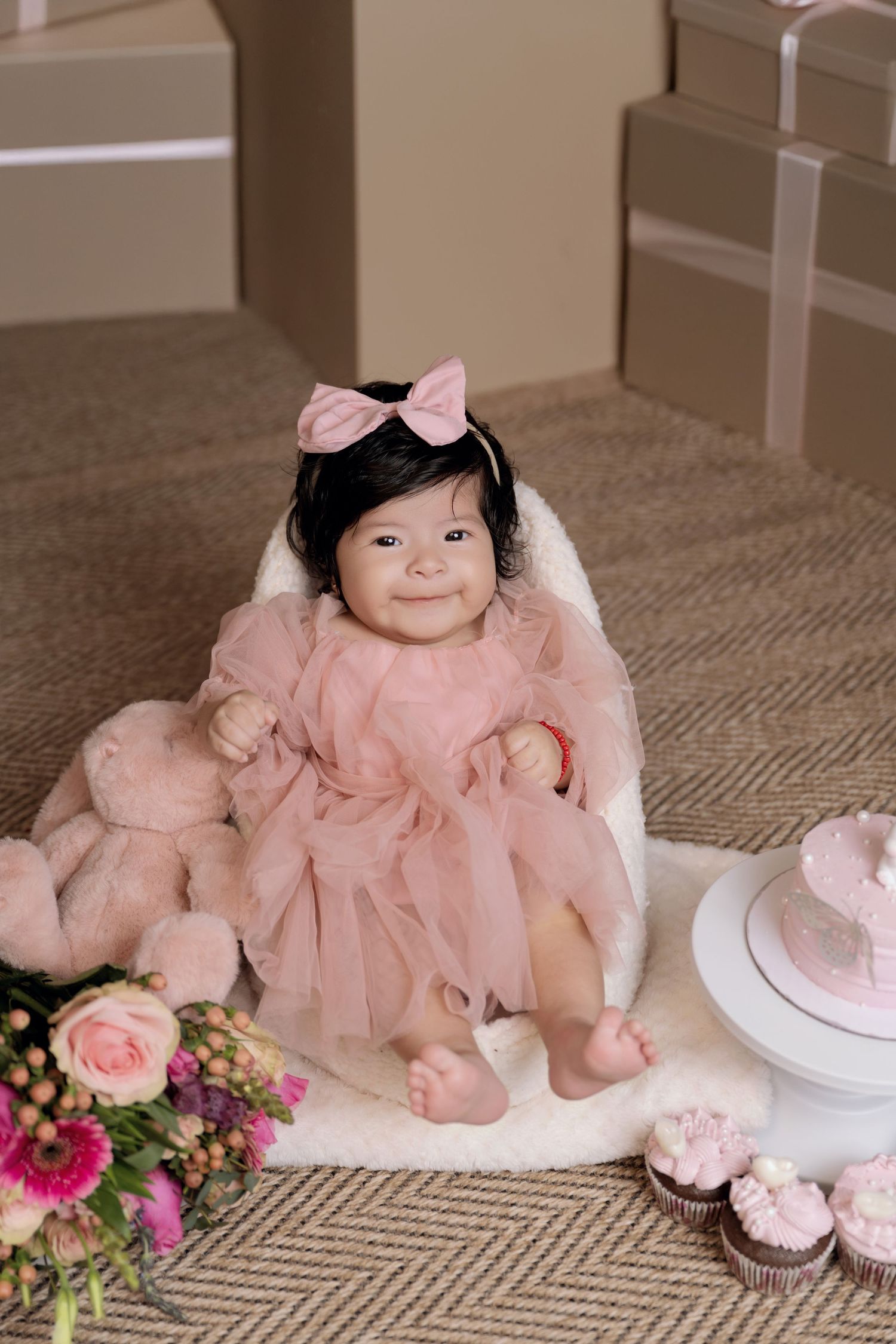 Smiling baby girl in pink tulle dress and bow headband sits near birthday cake, cupcakes, flowers, and gift boxes.