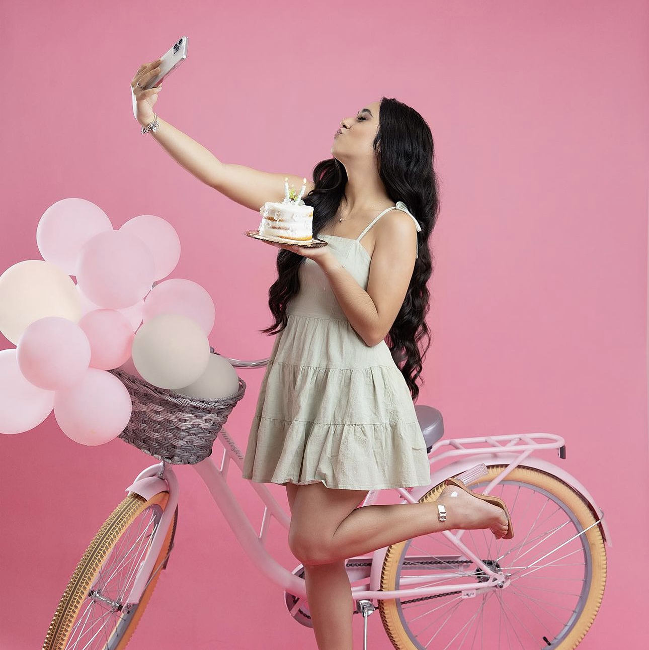 Young woman in white dress taking selfie while holding birthday cake beside pink bicycle with balloons on pink background.