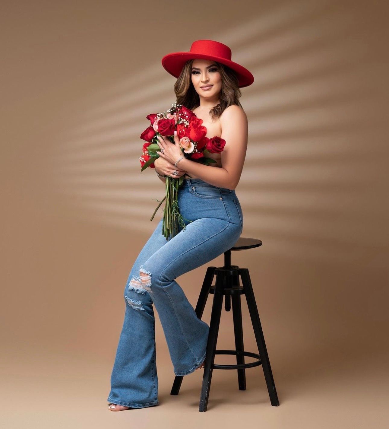 Woman in red hat and jeans holding red roses bouquet seated on stool for elegant portrait photo.