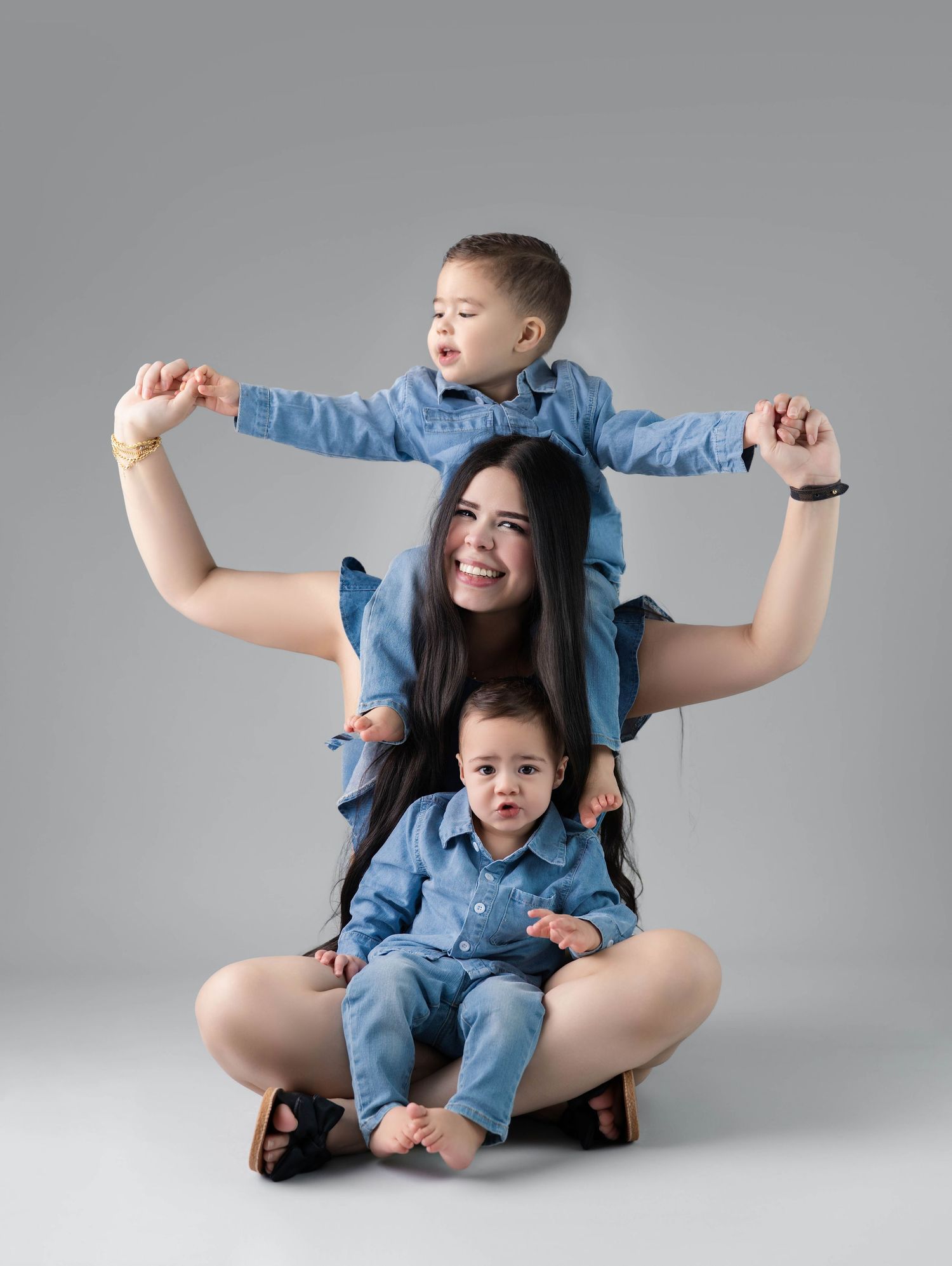 Mother sits cross-legged with toddler on shoulders and baby in lap, all wearing matching blue denim outfits.