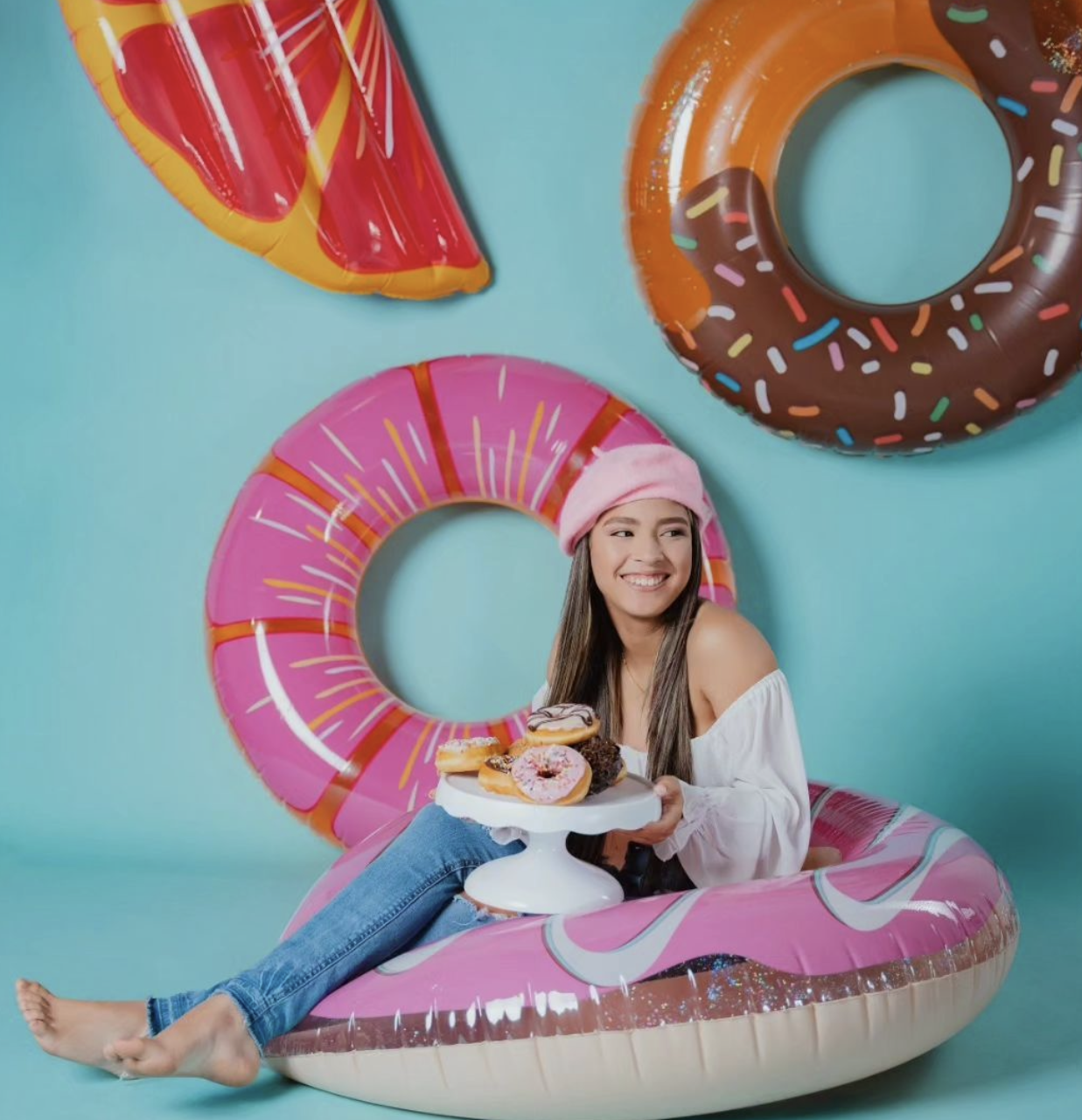 Young woman sitting in a pink donut pool float holding donuts, surrounded by inflatable food floats on blue background.