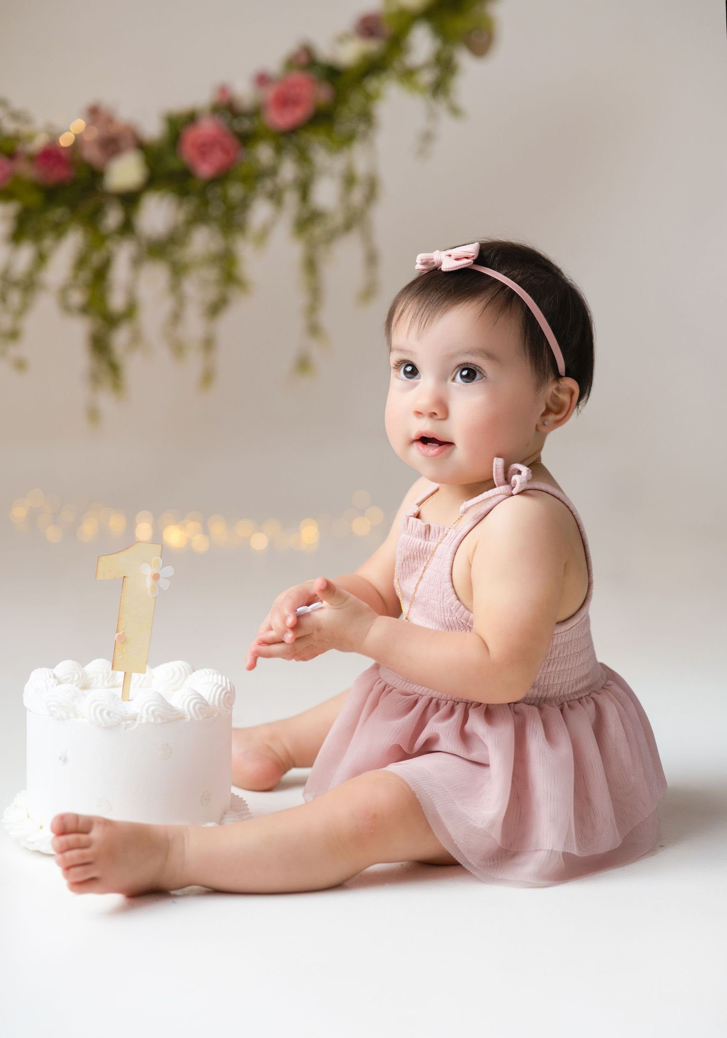 Baby girl in pink dress celebrates first birthday with white cake topped with gold number one topper, floral garland backdrop.