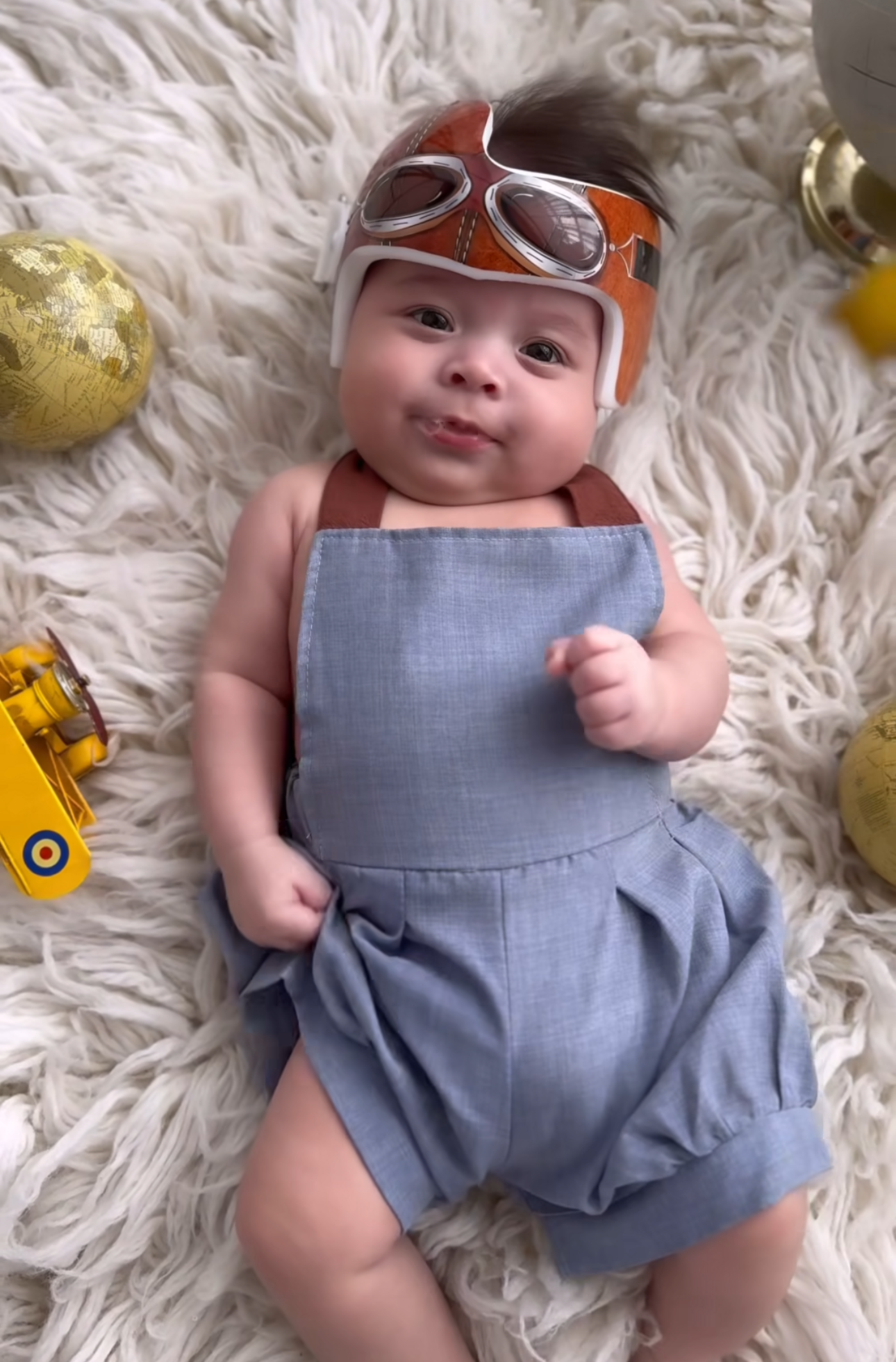 Adorable baby in a blue romper and aviator helmet with goggles lies on fluffy white rug with toy airplane.