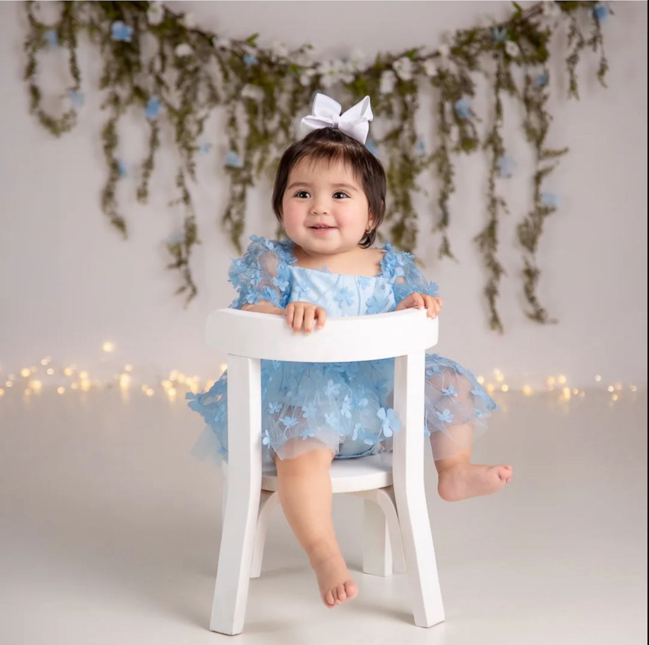 Baby in blue tulle dress sits on white stool against floral backdrop with twinkling lights.