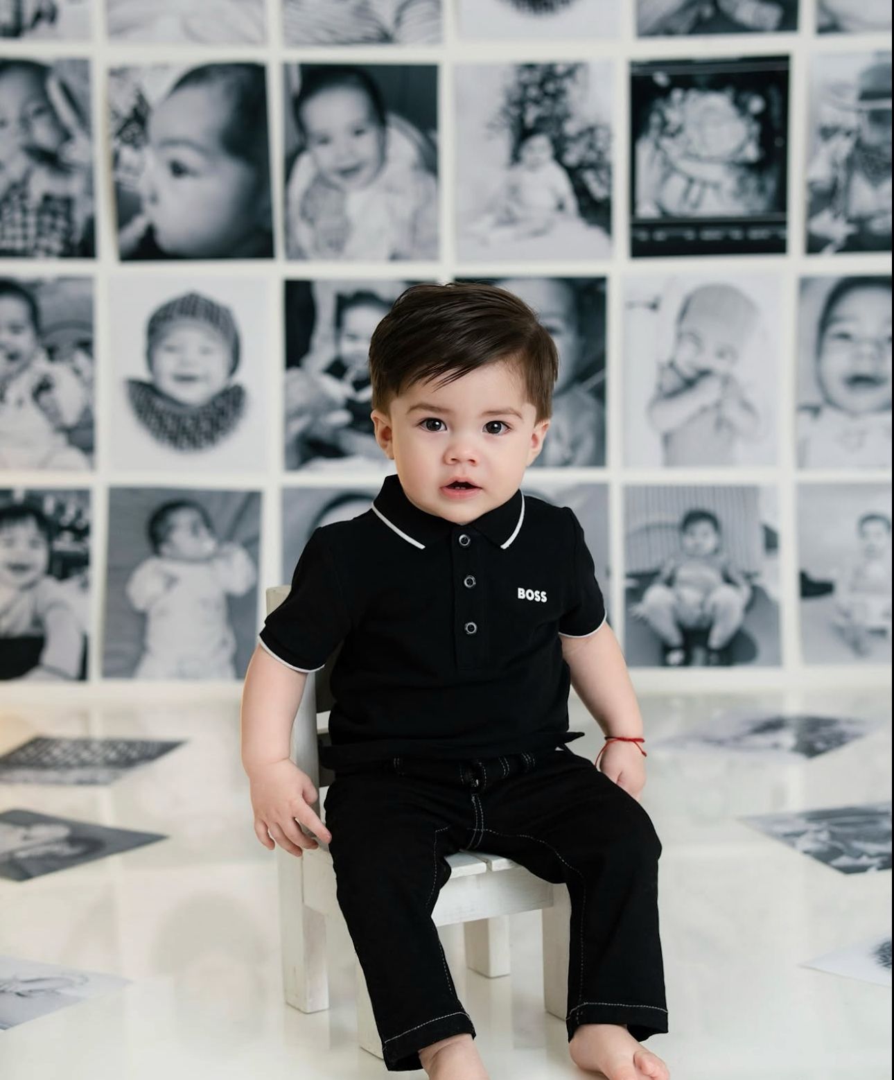 A young child in a black polo outfit sits on a white chair against a wall of vintage baby photographs.