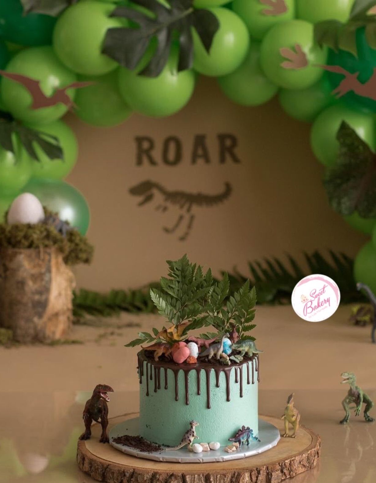 Dinosaur themed birthday party setup with green balloon arch and ROAR sign on tan wall.