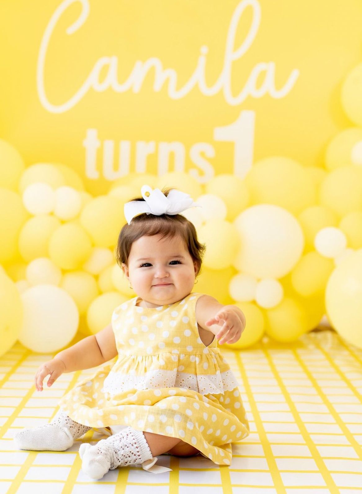 Baby in yellow polka dot dress sits on yellow backdrop with white text overlay for first birthday celebration.