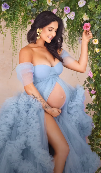 Maternity photoshoot with flowing blue tulle gown against floral backdrop showing expectant mother in elegant poses.