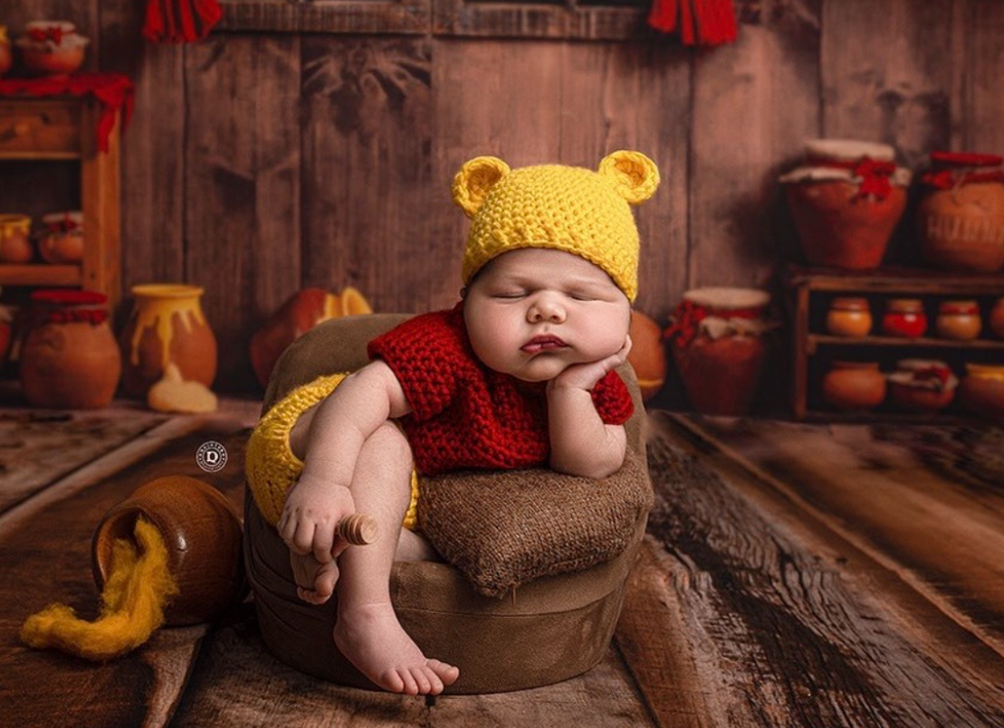 A sleeping baby wearing a yellow knit hat lies peacefully on a brown blanket with rustic wooden backdrop and pottery decor.