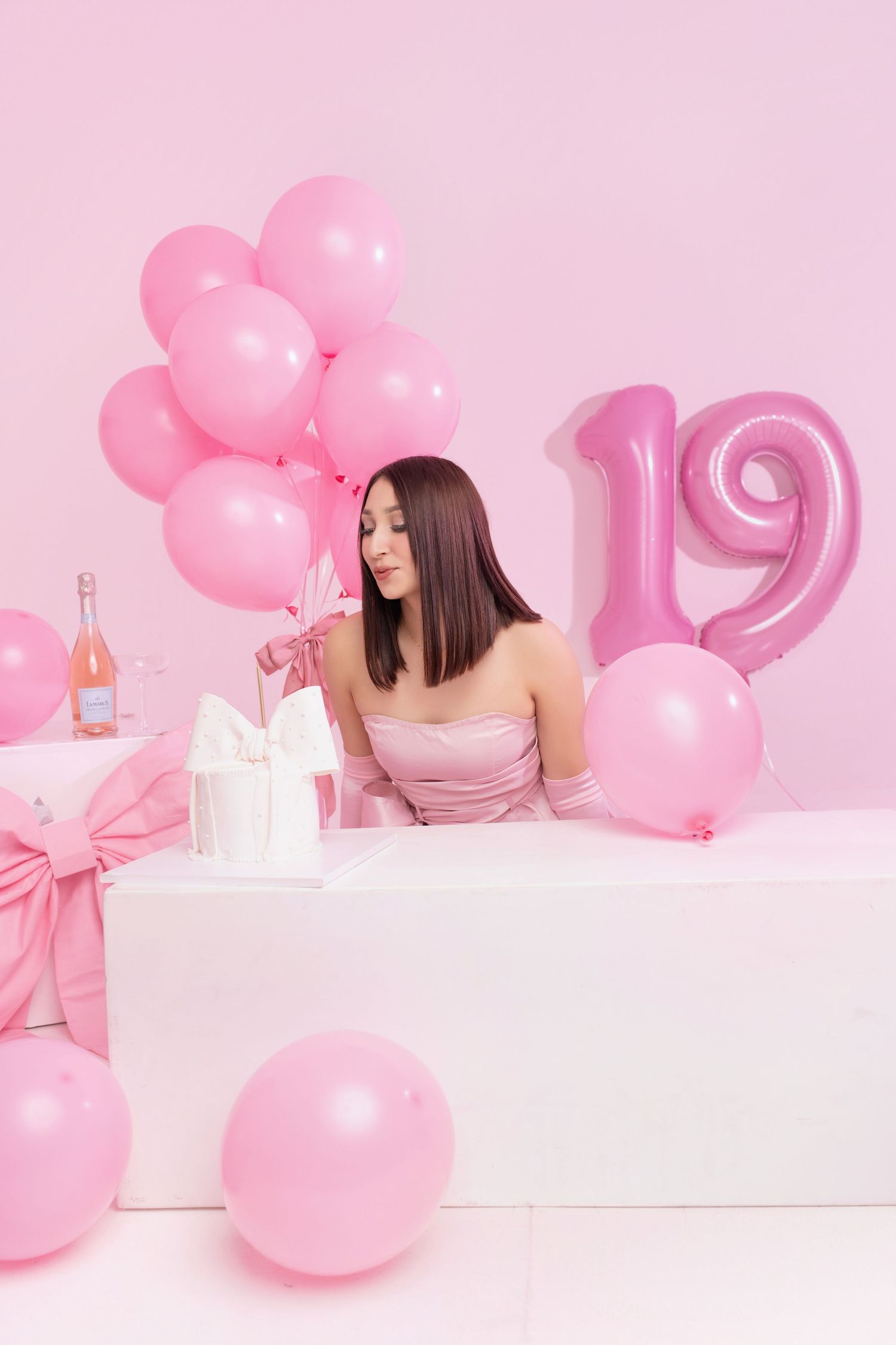 A pink monochromatic photoshoot setup with pink balloons, white table, and a person in pink dress posing elegantly.