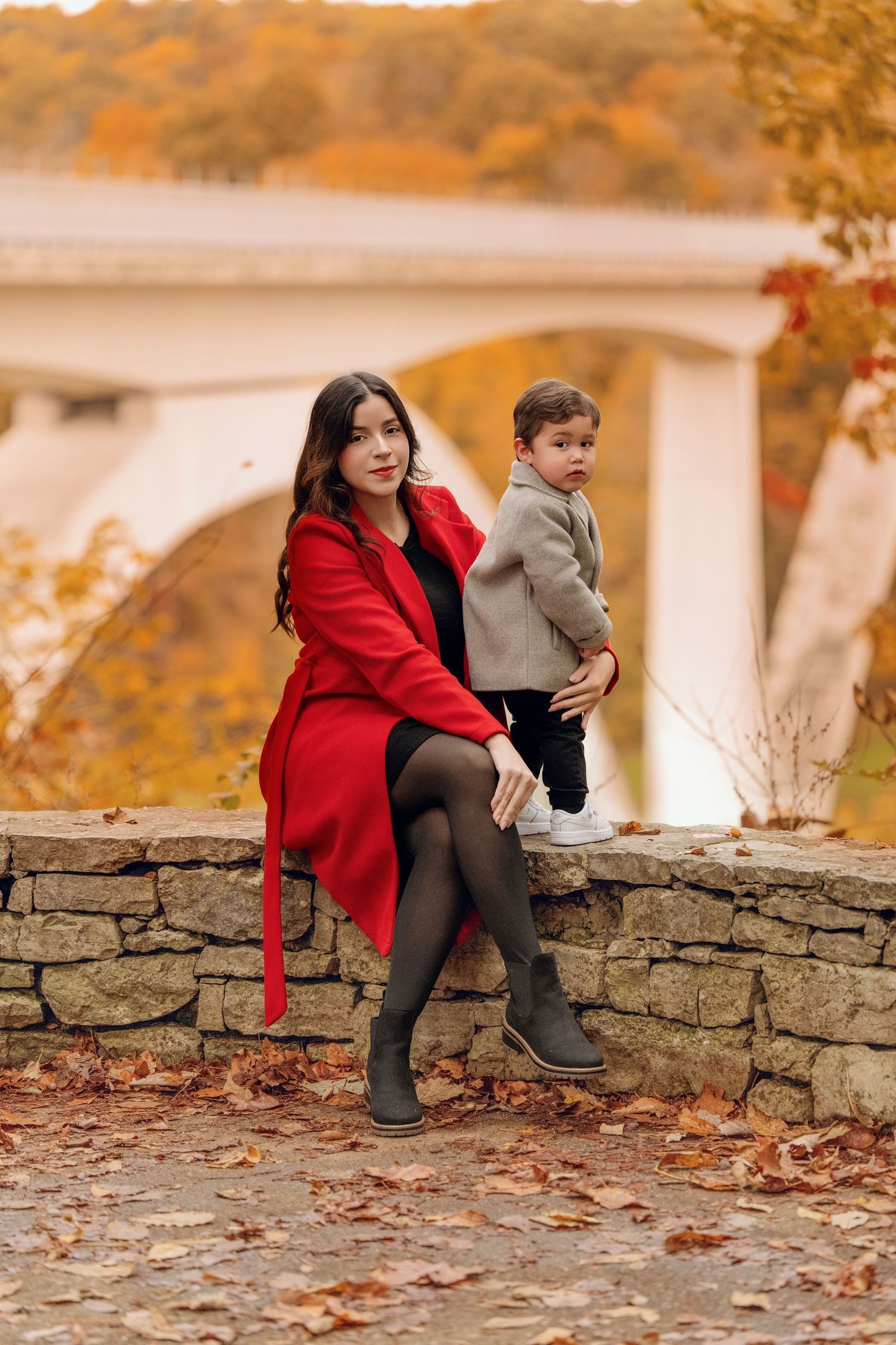 A woman in a red coat sits on a stone wall holding a toddler boy amid colorful autumn foliage near a bridge.