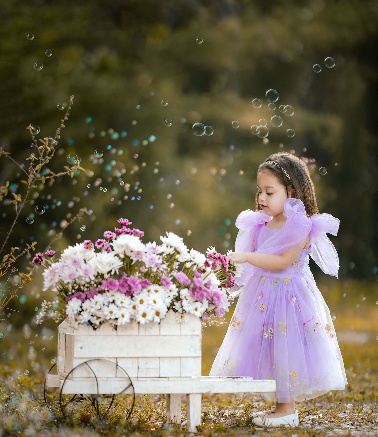 Little girl in purple tulle dress touches flowers in white cart surrounded by floating bubbles outdoors.