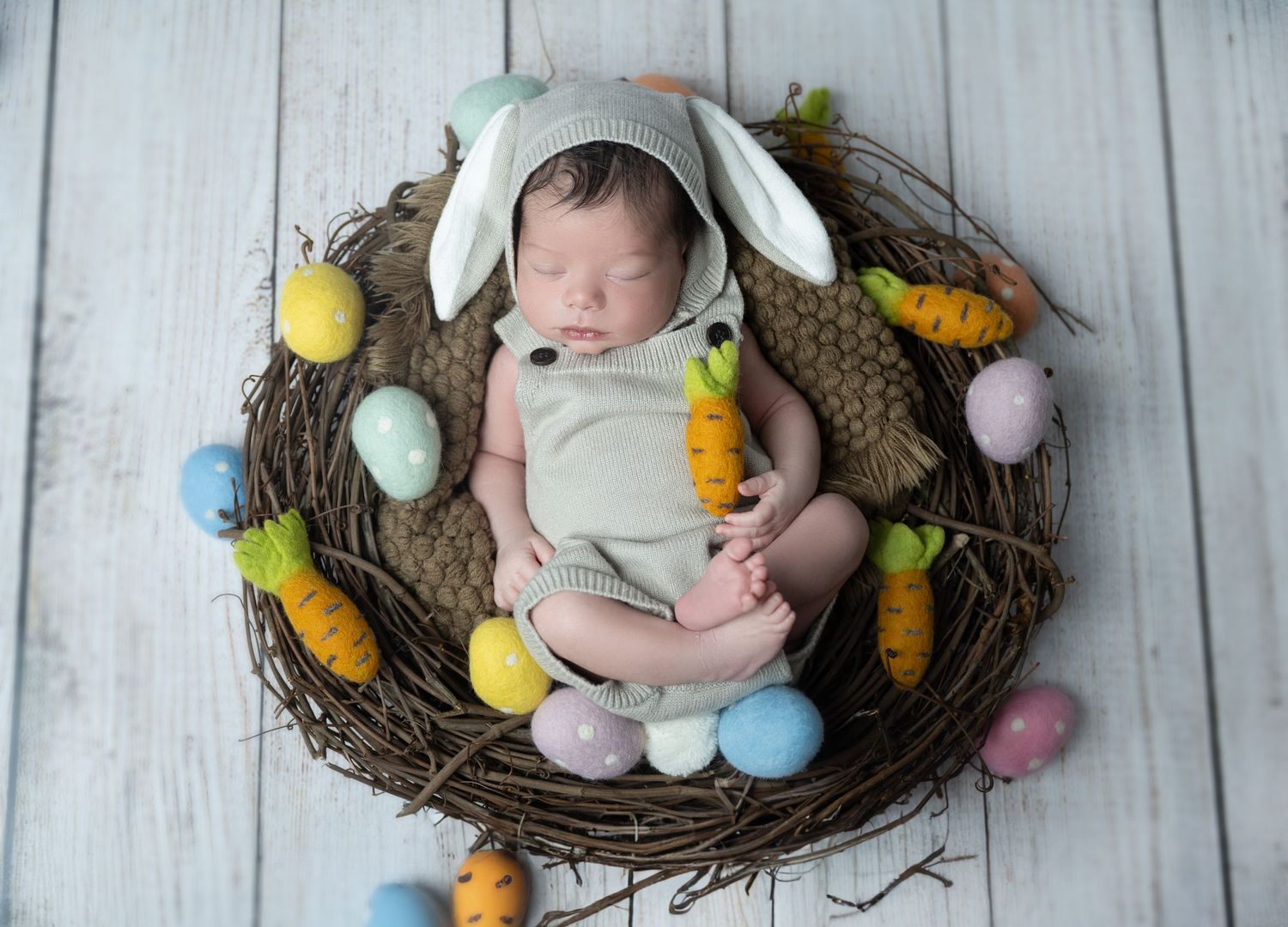 Sleeping newborn in bunny outfit nestled in Easter egg decorated nest.