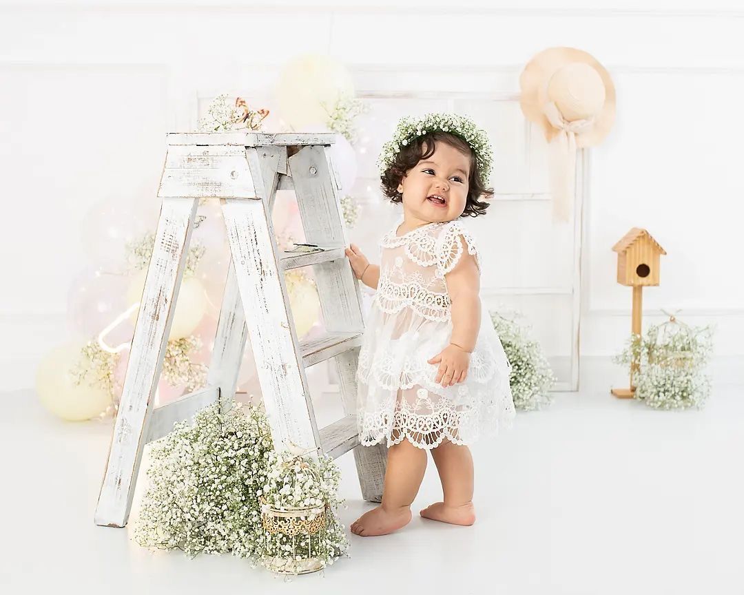 Baby girl in white lace dress with floral crown stands by rustic ladder with balloons and flowers in bright studio.