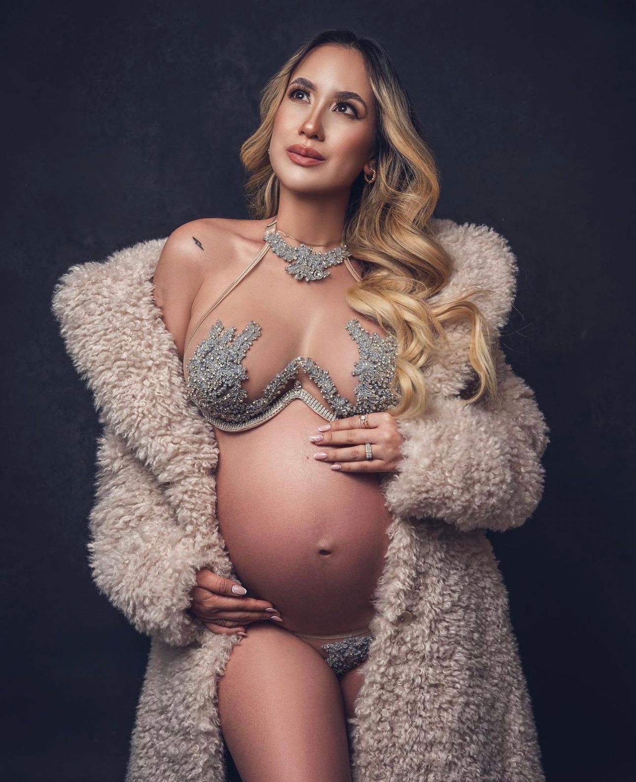 Pregnant woman in jeweled bra and matching underwear, wearing a fluffy beige coat, posing for a maternity photoshoot.