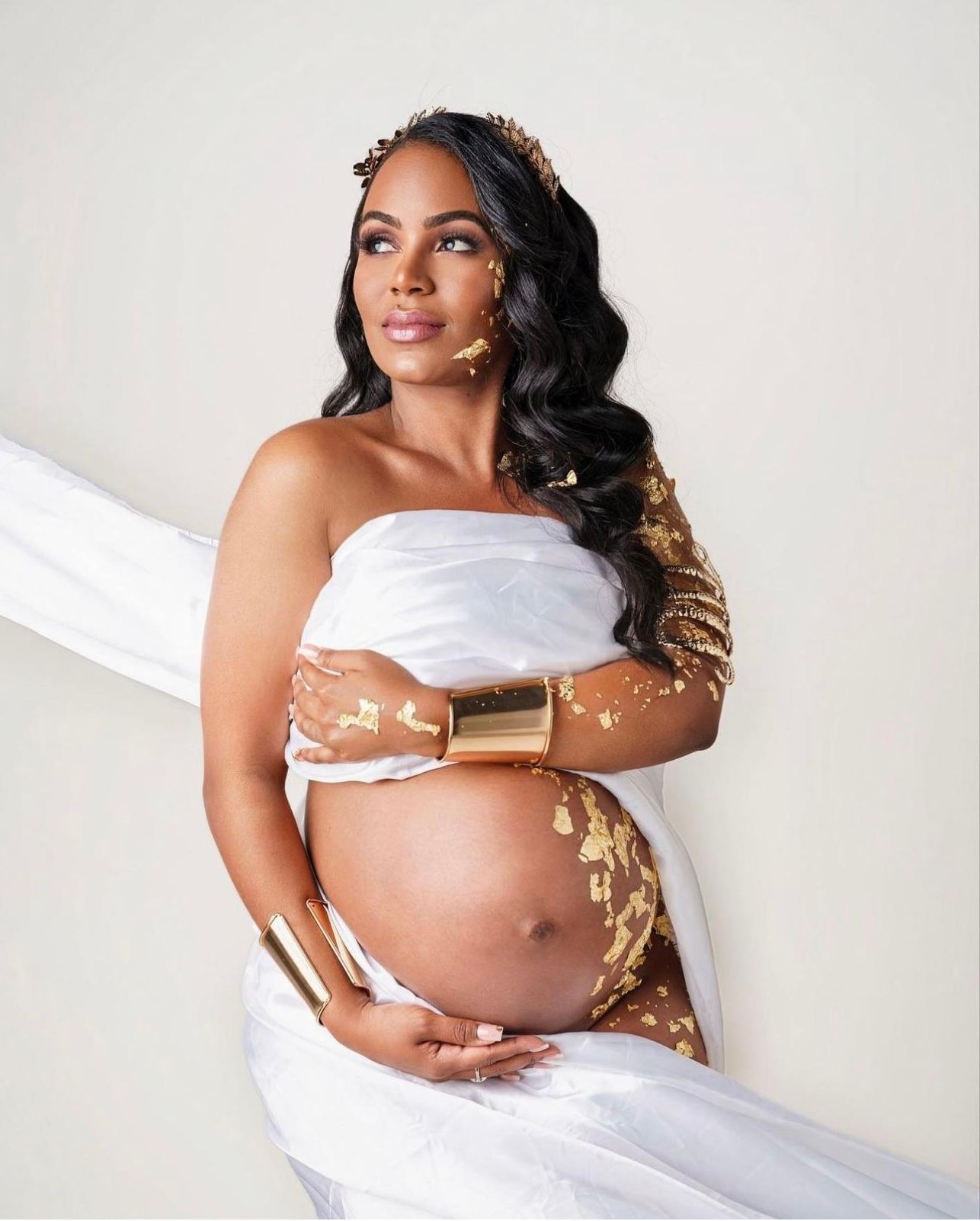 Pregnant woman in elegant white draped fabric with gold accessories and crown posing for a maternity photoshoot.