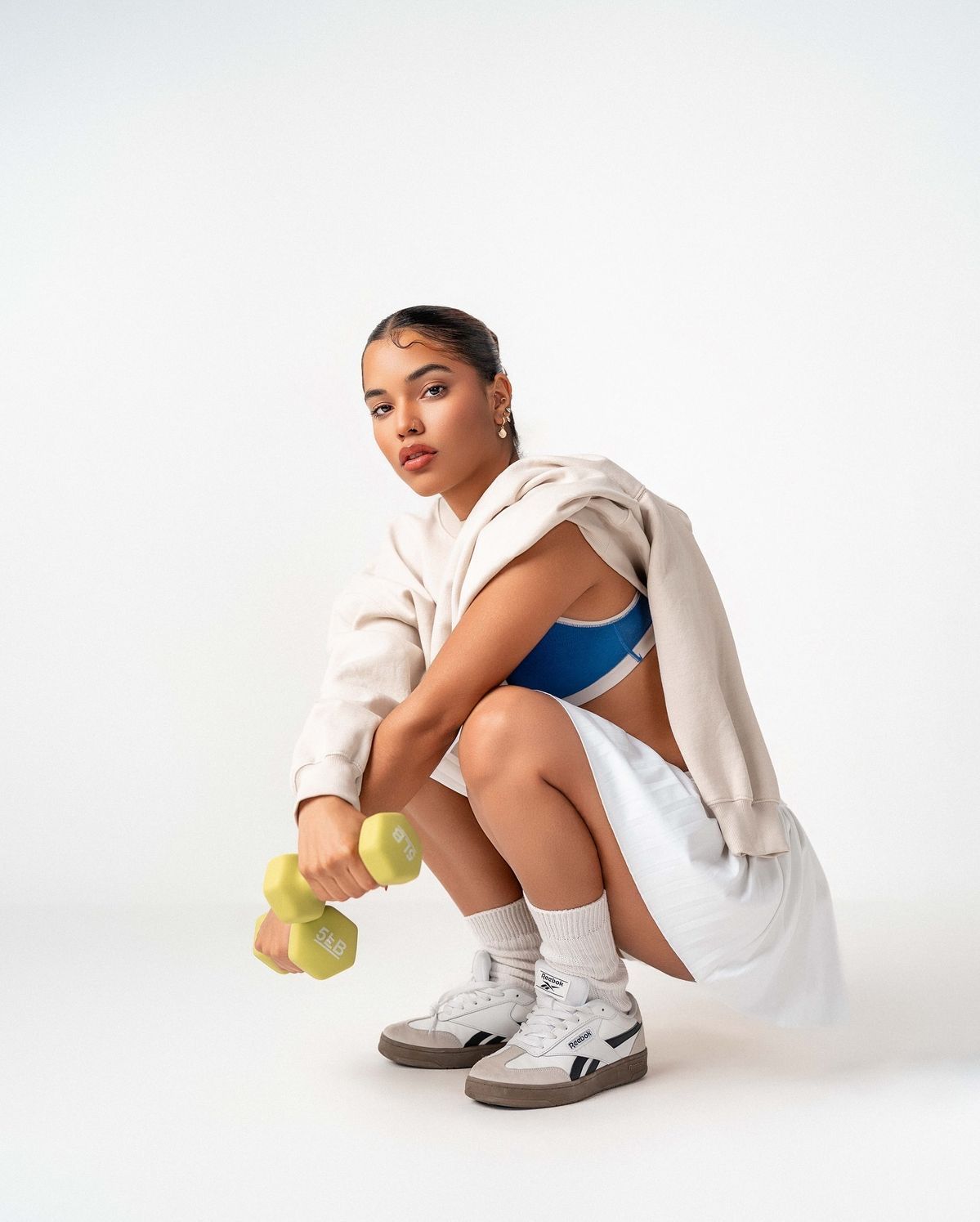 Woman crouching in blue sports bra, white shorts, hoodie, and Reebok sneakers, holding green dumbbells.