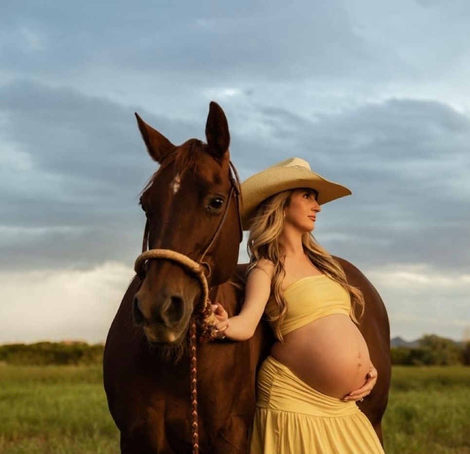 Pregnant woman in yellow dress and cowboy hat stands beside a brown horse in a field under a cloudy sky.