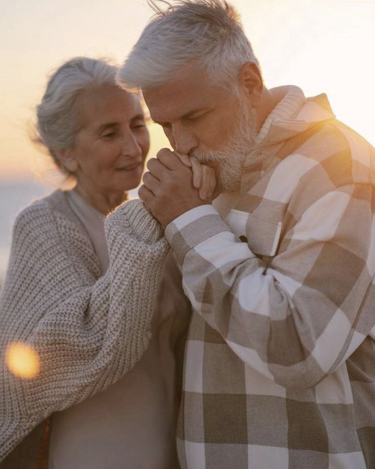 Elderly couple sharing a romantic moment at sunset, man kissing woman's hands tenderly outdoors.