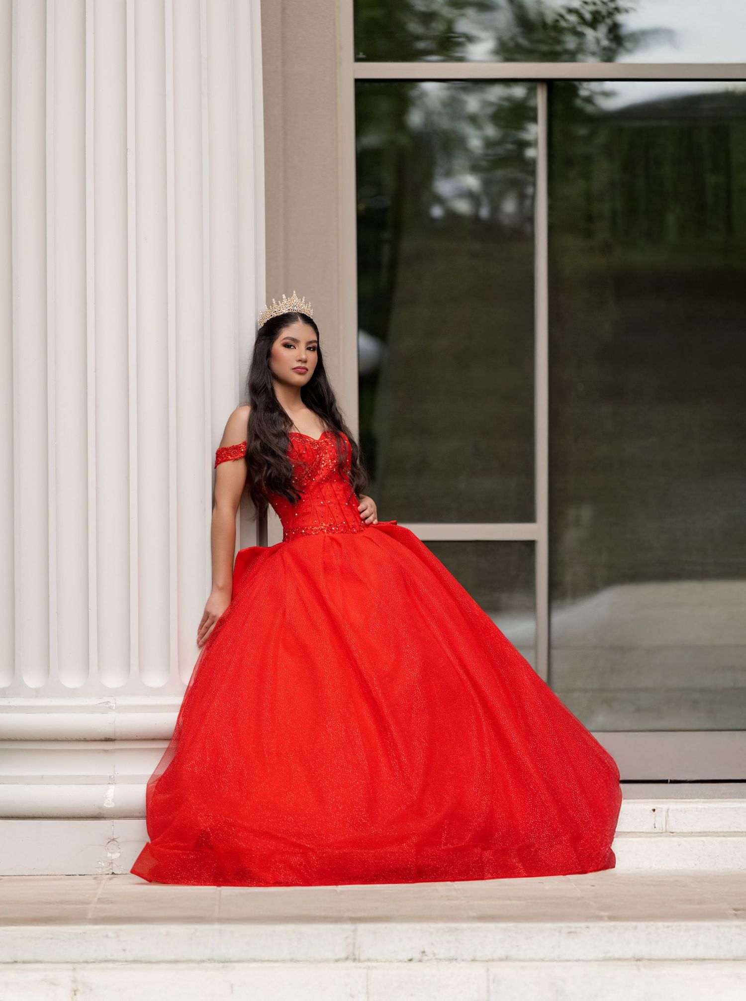 Young woman in red off-shoulder ball gown with gold crown poses on marble steps for quinceañera photos.