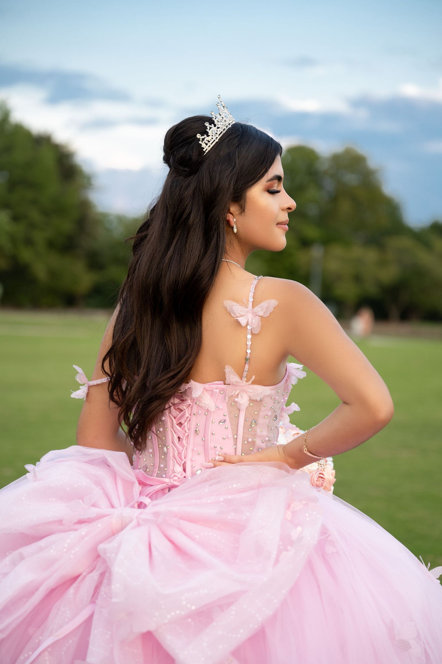 Young woman in pink quinceañera ball gown with butterfly details and tiara poses outdoors on green grass.