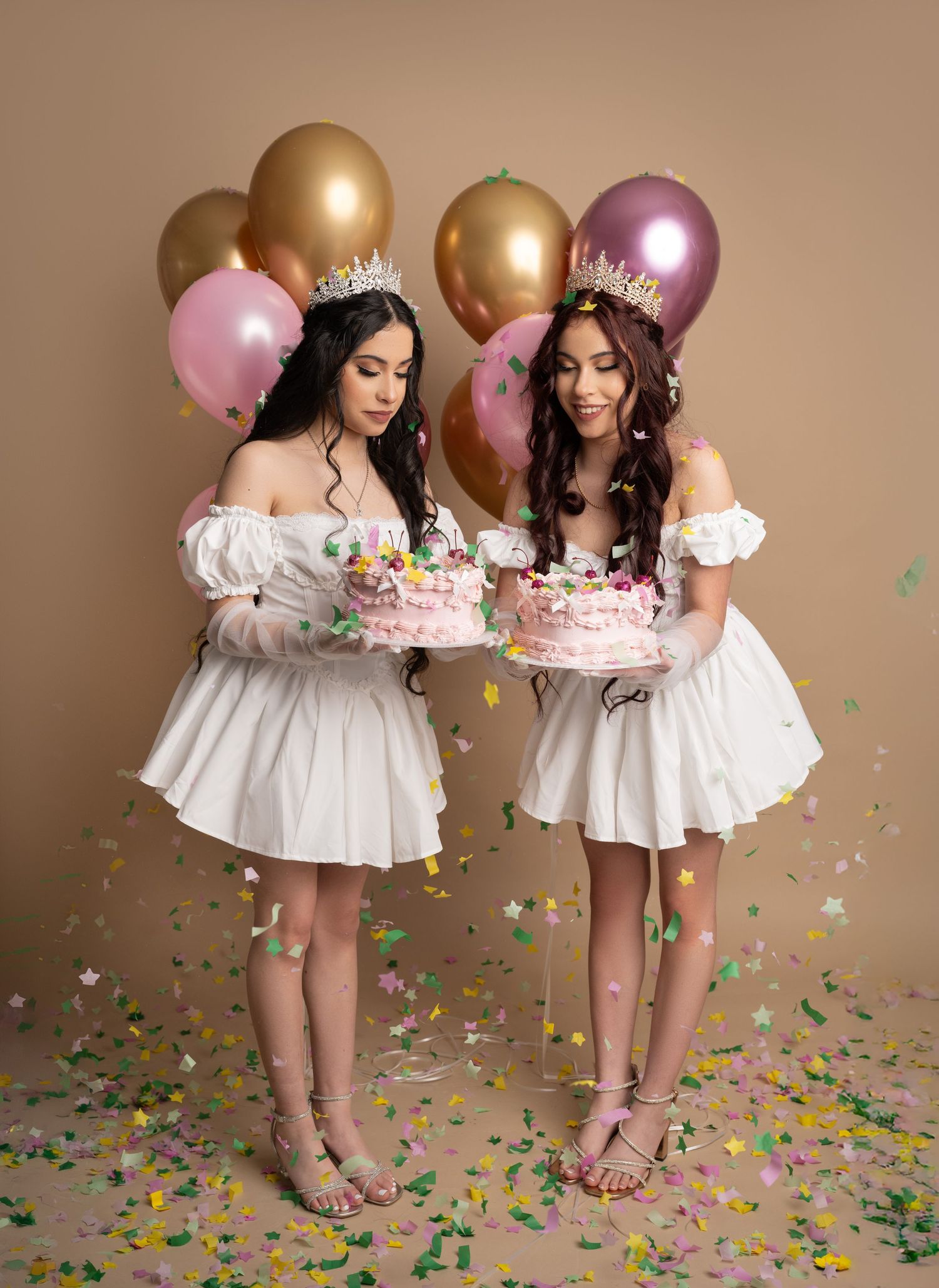 Two young women in white dresses and gold crowns hold birthday cakes surrounded by colorful balloons and confetti.
