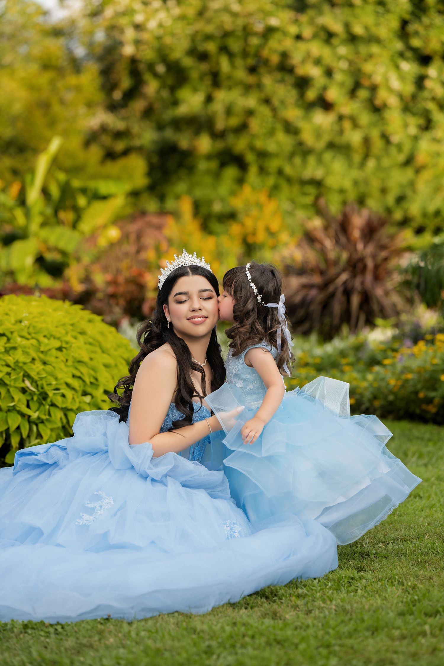 Two girls in matching blue ball gowns and tiaras share a sweet moment in a lush garden setting.