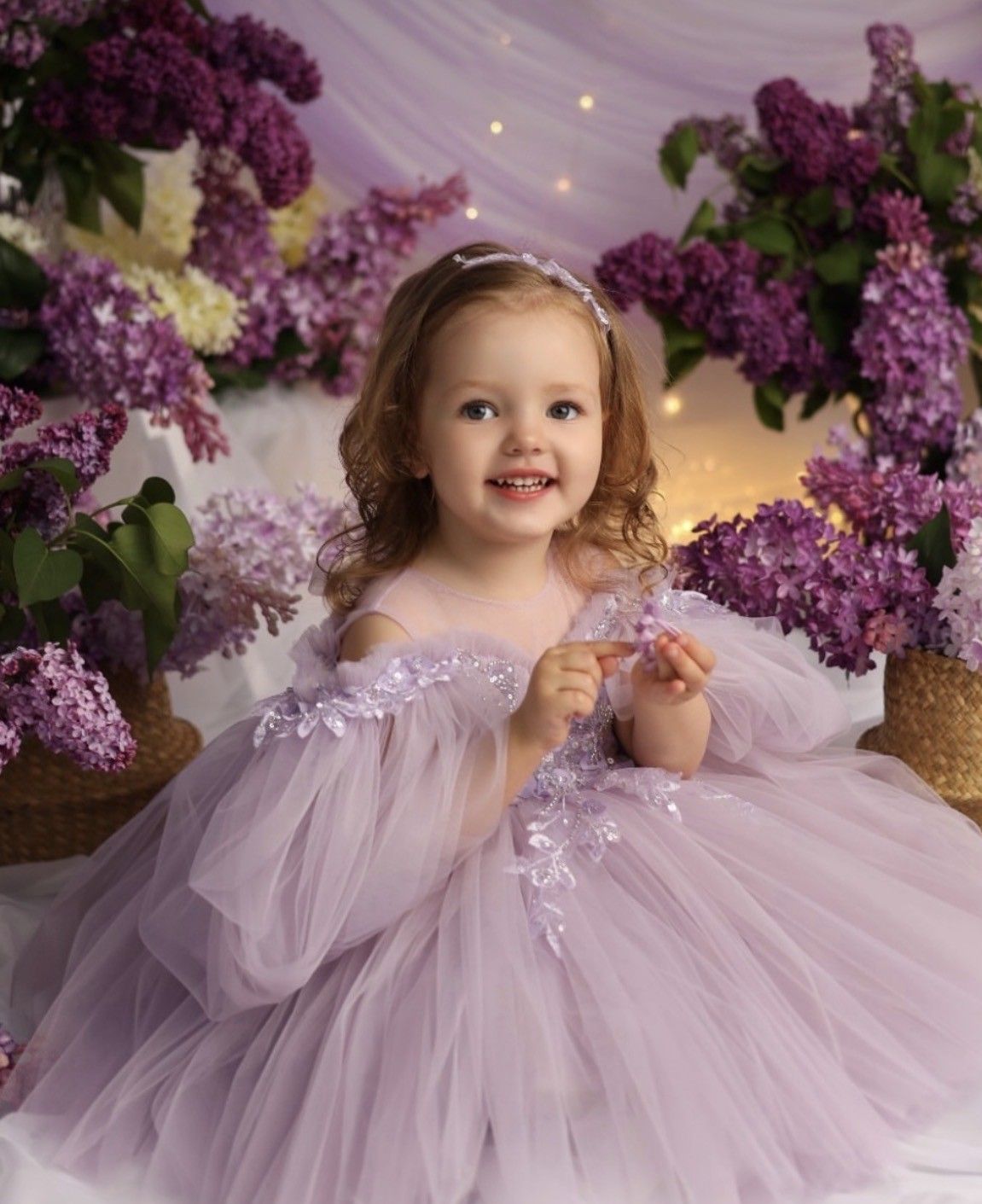 A smiling toddler girl in a lavender tulle dress poses among purple lilac flowers in a dreamy floral setting.