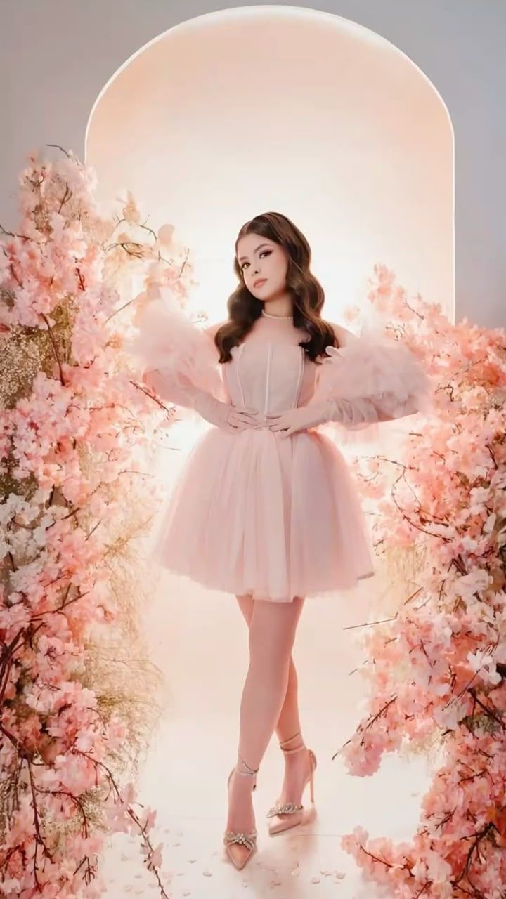 Young woman in pink tulle dress with puff sleeves poses among cherry blossom flowers in elegant studio setting.