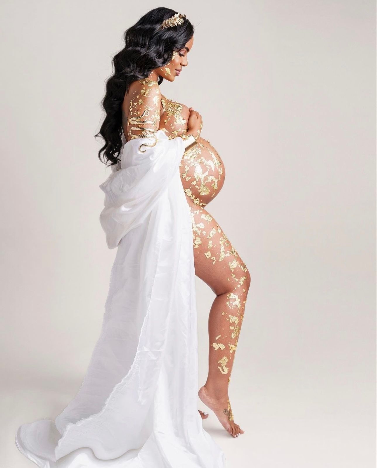 Pregnant woman in elegant white drape with gold floral body paint, posing sideways for a maternity photoshoot.