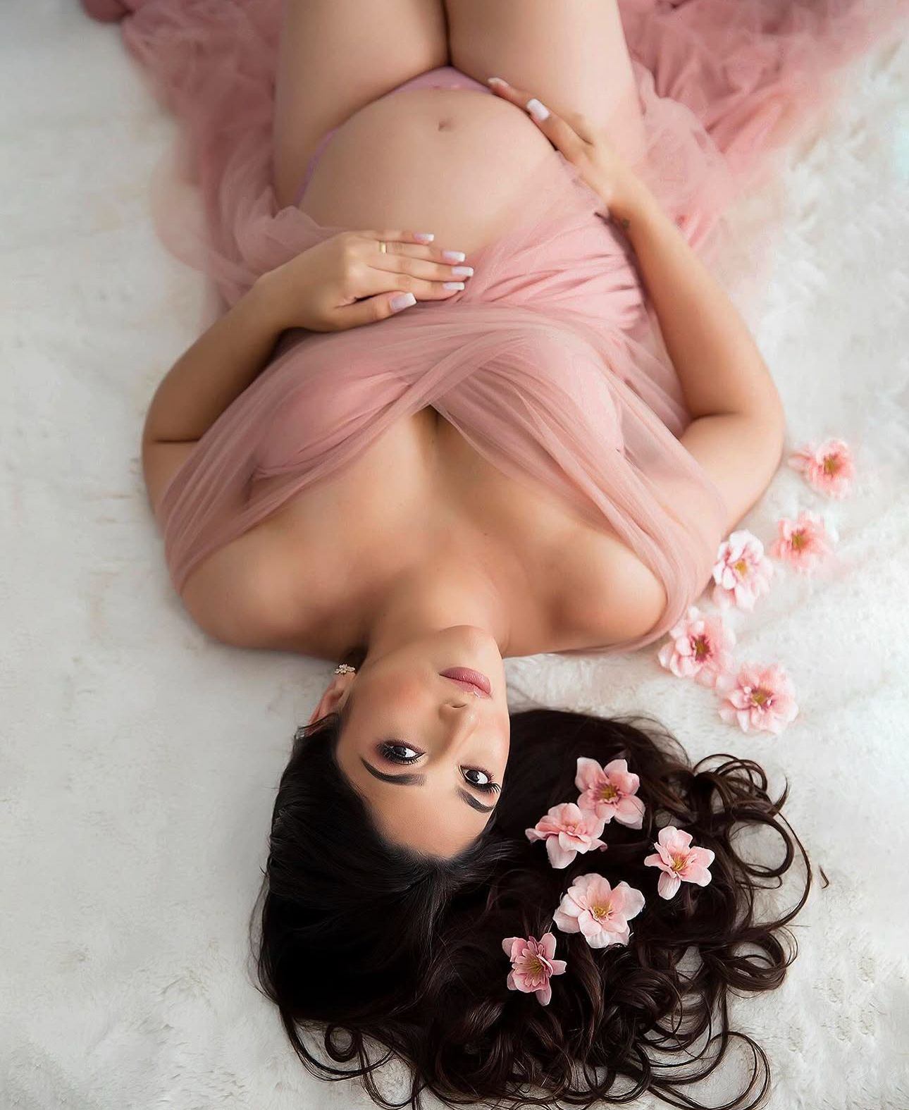 Pregnant woman lying down draped in pink tulle with flowers in her dark hair for a maternity photo shoot.