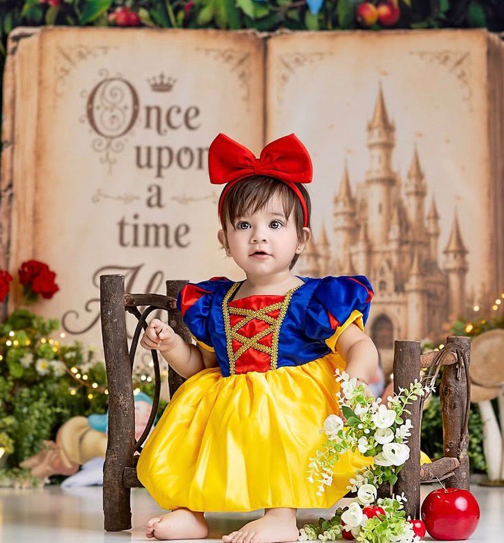 Toddler girl in Snow White costume with red bow headband sits on rustic bench before fairy tale backdrop.